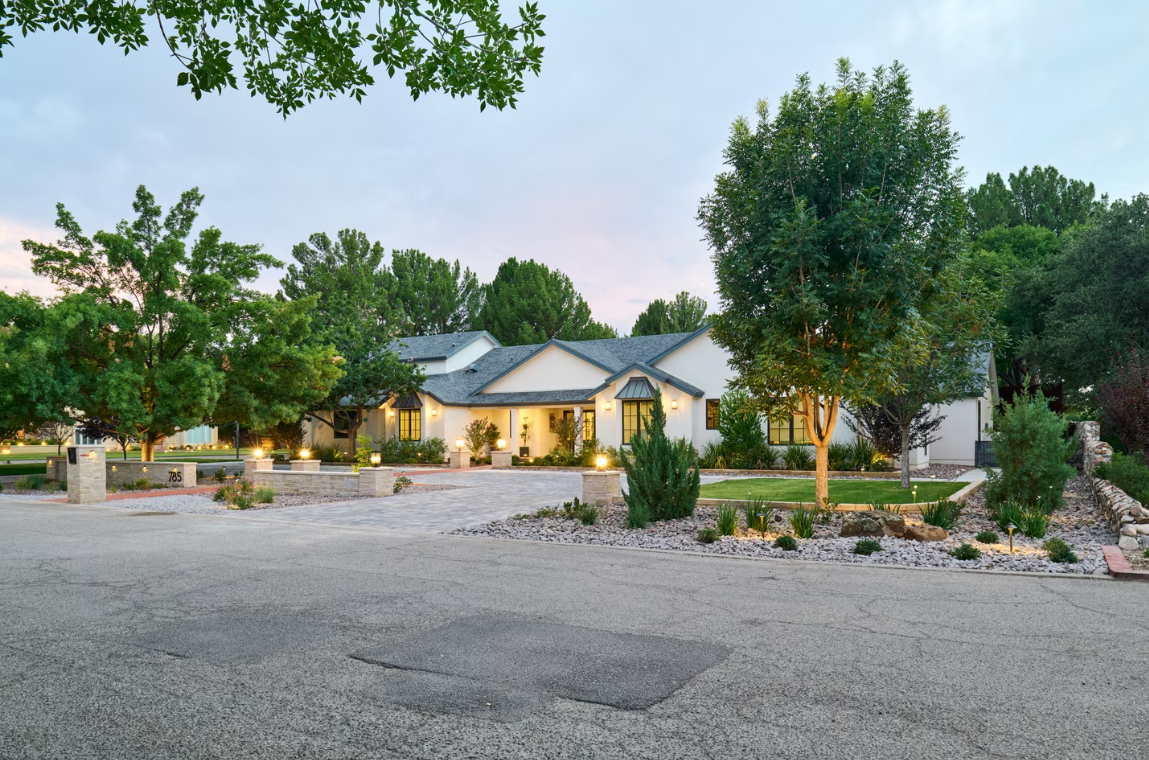 White house with gravel driveway and lush landscaping, lit with outdoor lights.