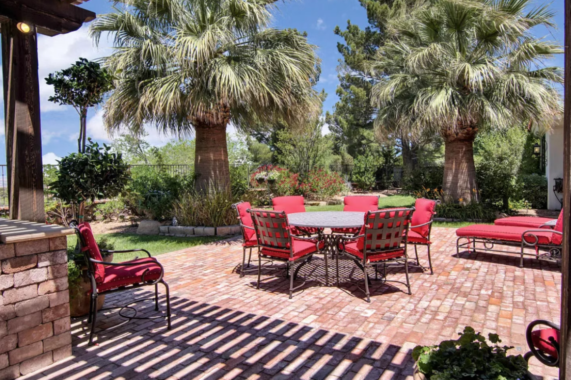 Patio with red chairs, round table, and lounge chairs on brick, under palm trees.