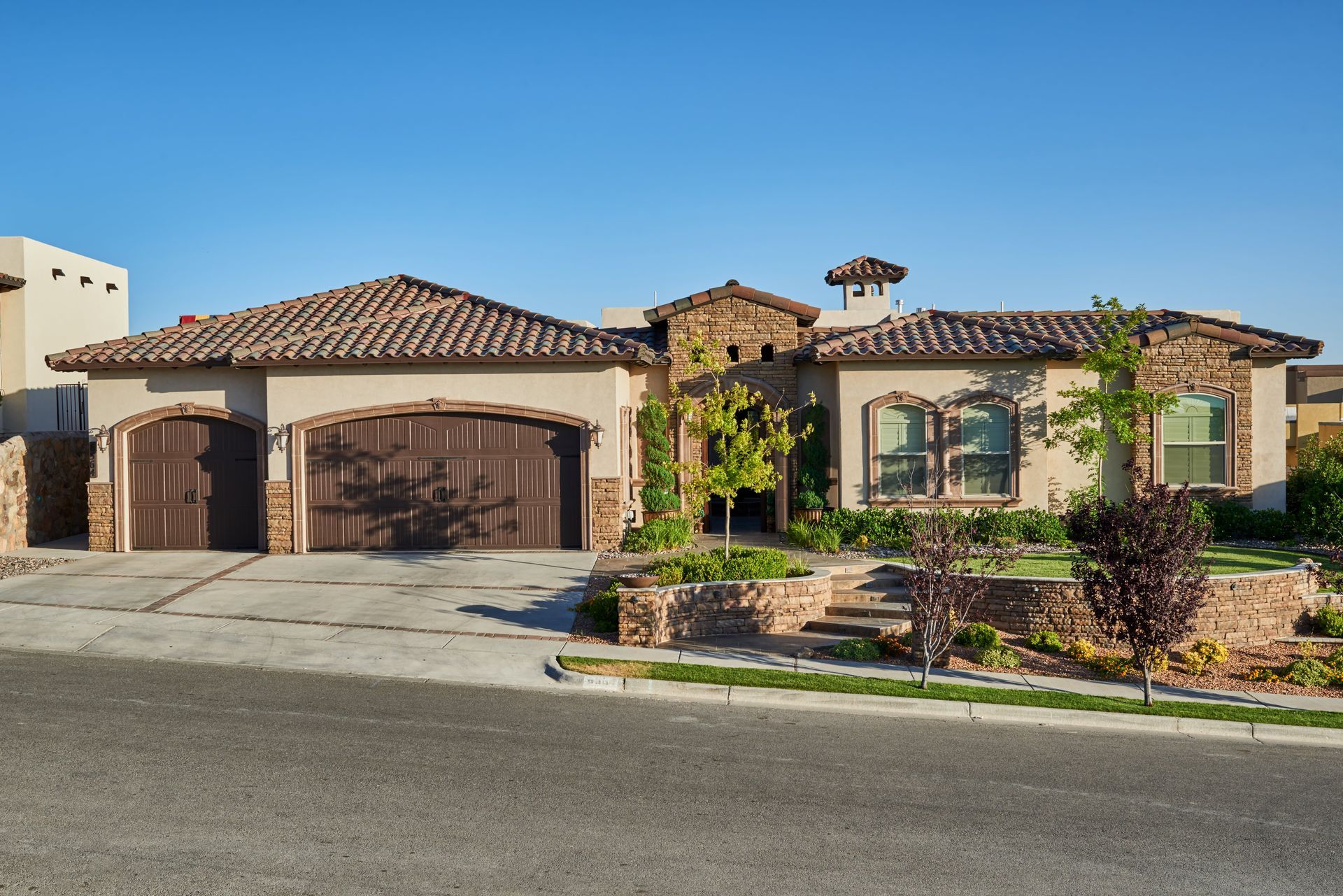 Beige stucco house with brown tiled roof, wooden garage doors, and landscaped front yard.