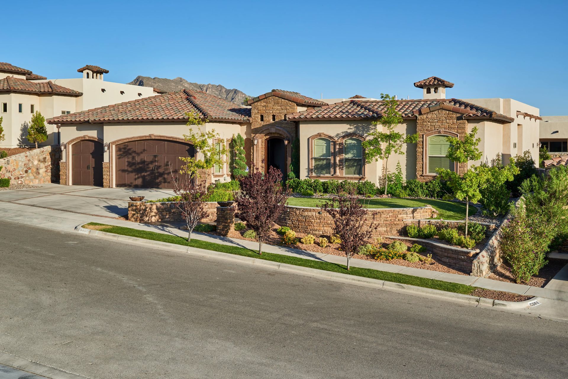 House with stone facade, tile roof, and attached garage on a sloped street under a blue sky.