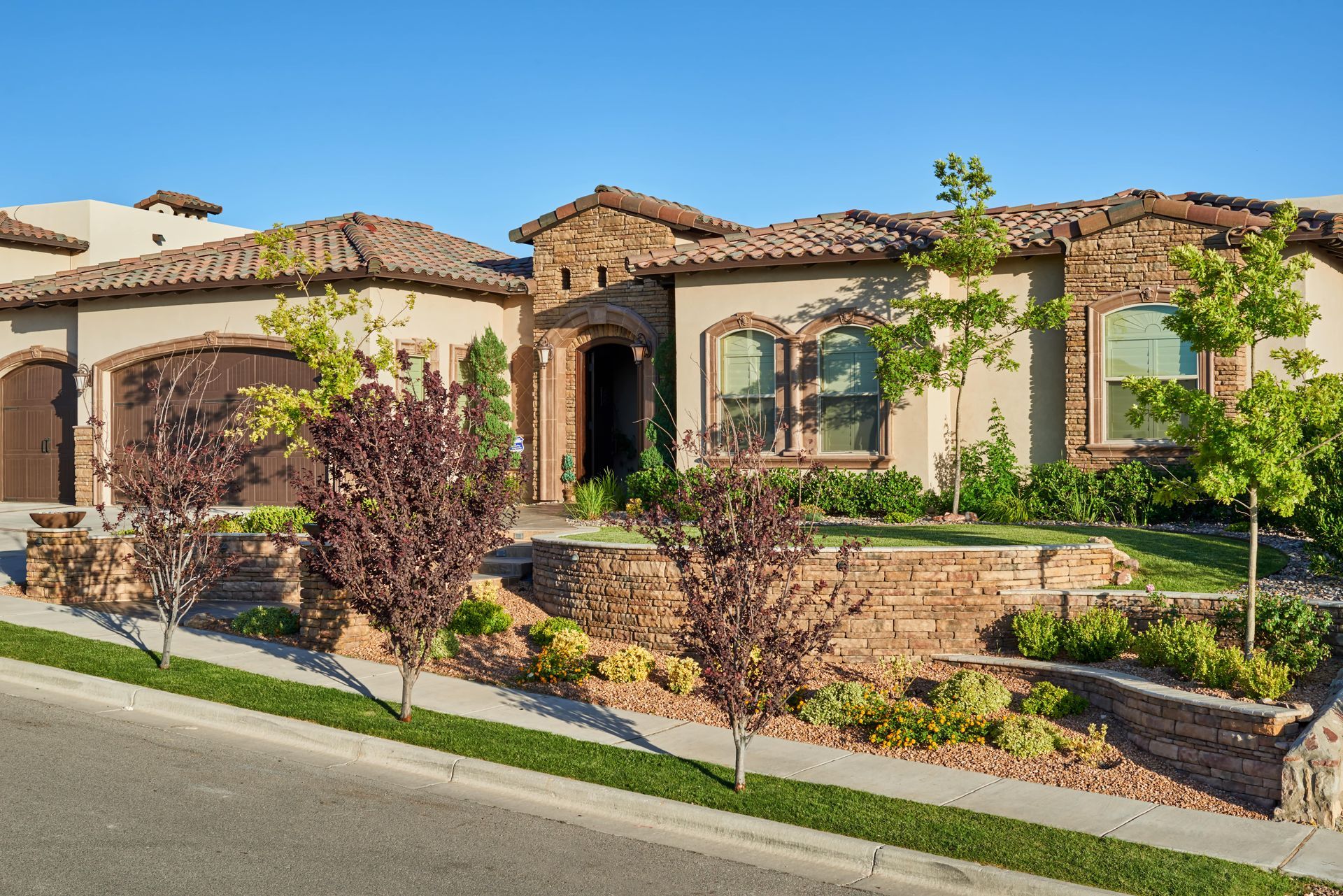 A beige and brown brick house with a tile roof and tiered landscaping under a blue sky.