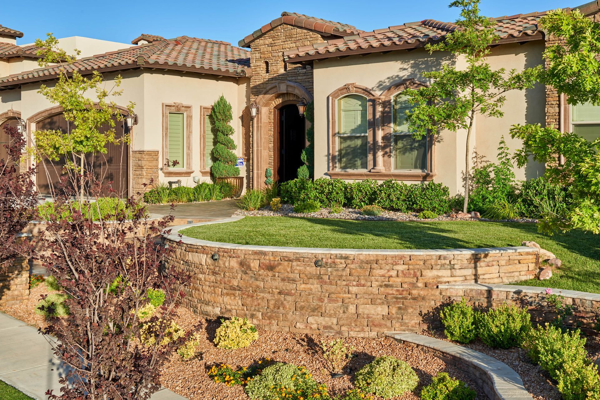 Beige stucco home with stone accents, terracotta roof, and terraced landscaping with green grass and shrubs.