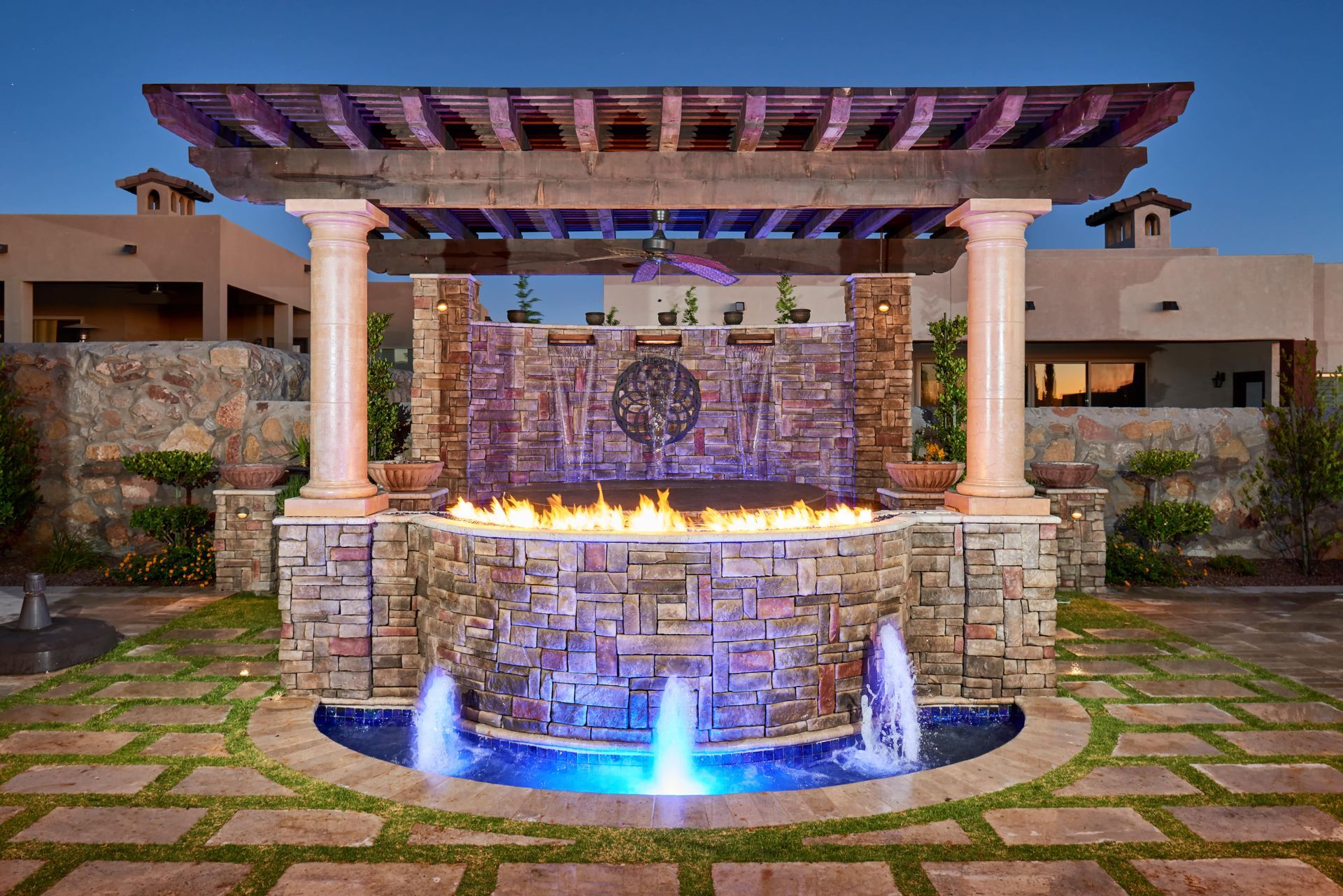 Stone water fountain with fire feature, under wooden pergola, illuminated blue and yellow.