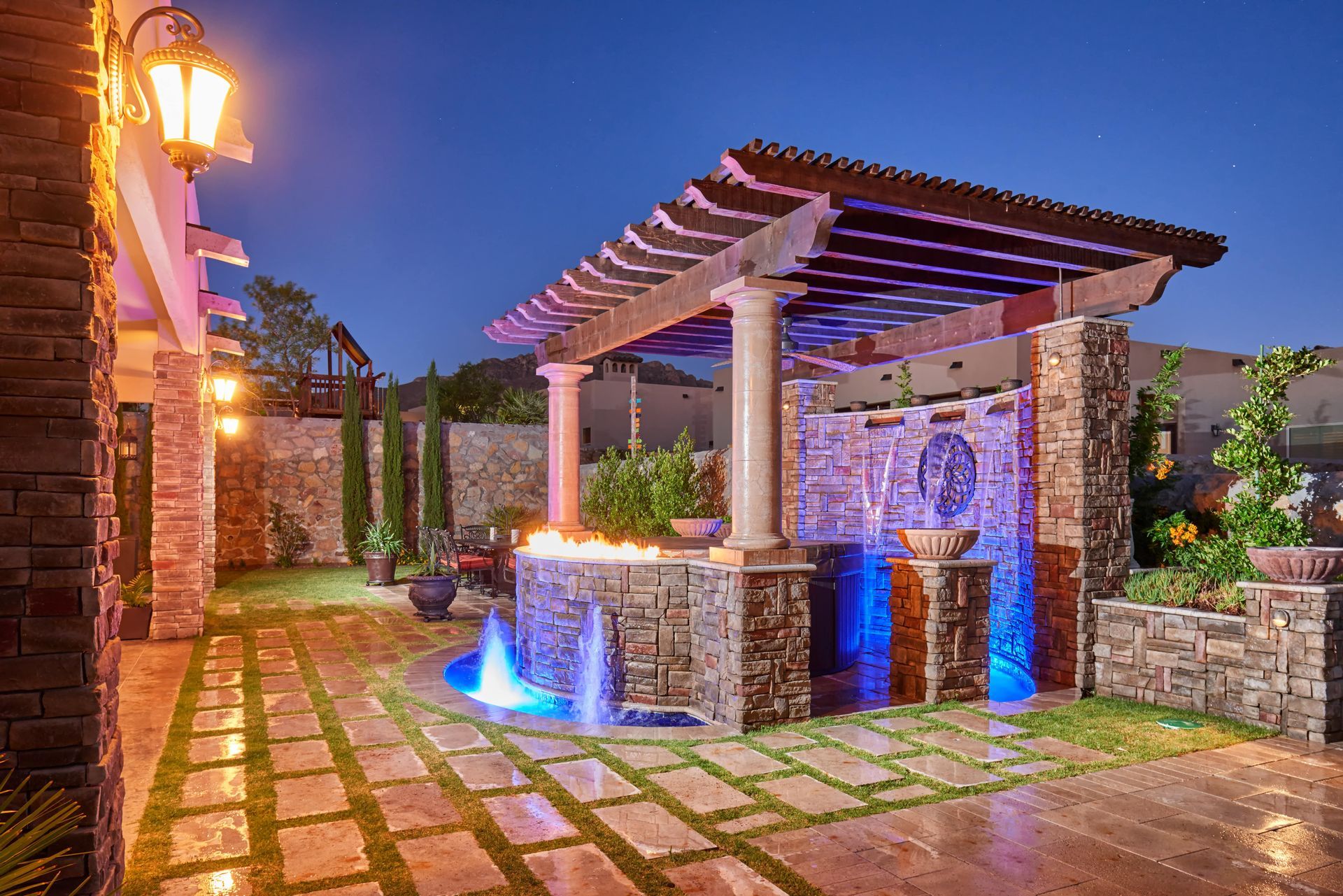 Outdoor oasis with a stone water feature, pergola, and blue-lit water jets at dusk.