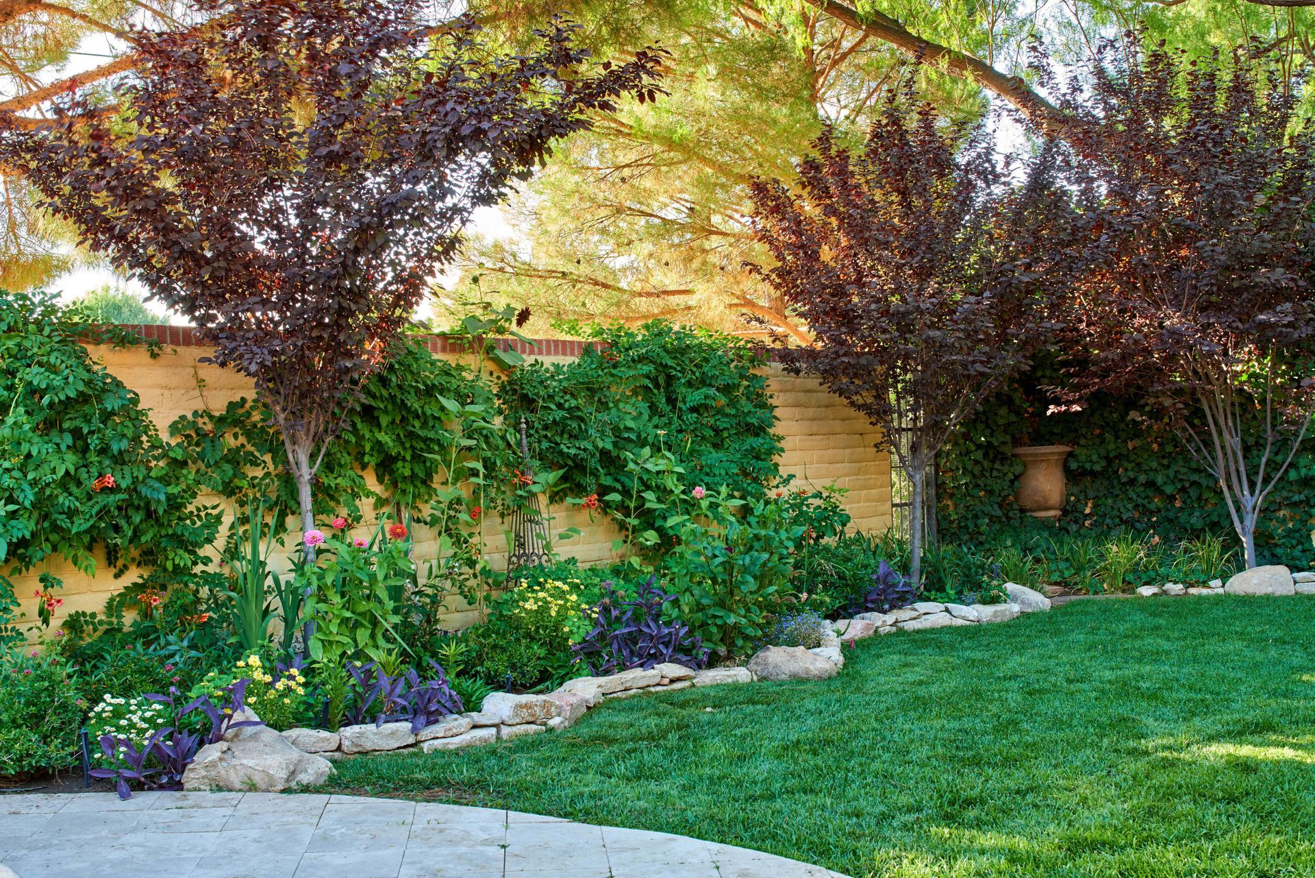 Lush garden with a stone-lined border, vibrant green grass, and flowering plants against a light-colored wall.