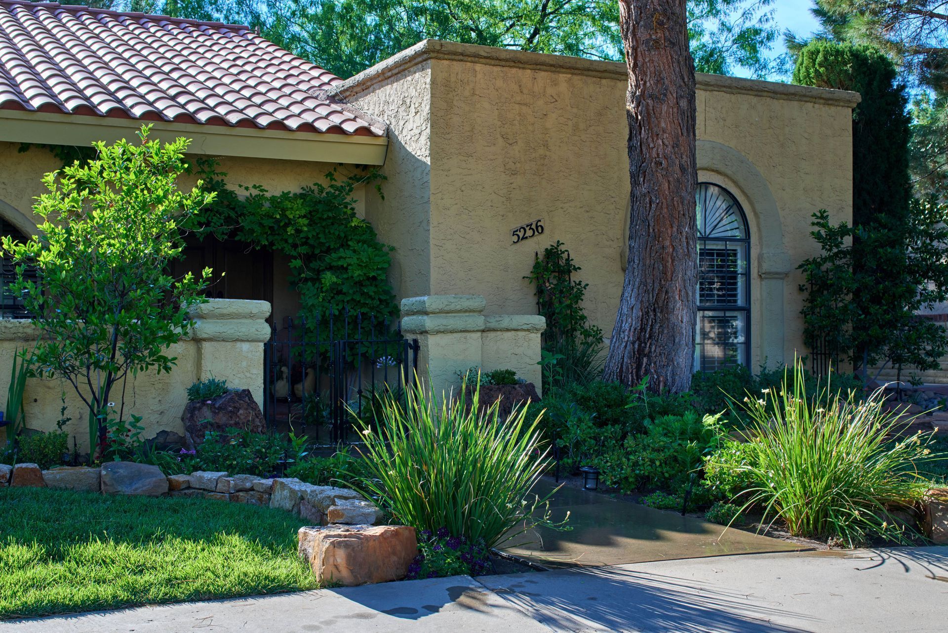 Beige stucco house with a tiled roof, lush landscaping, and a large tree in front.
