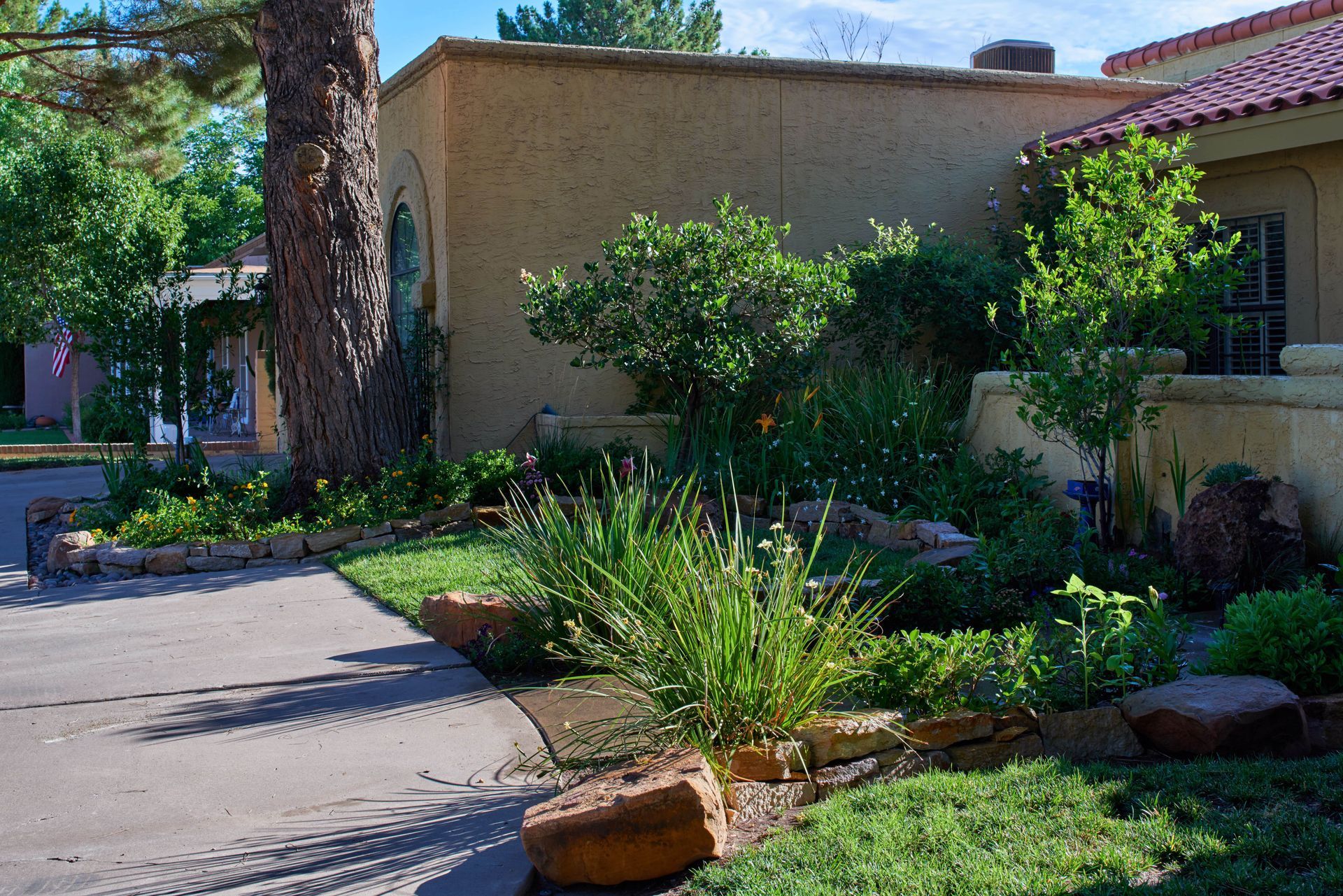 Sidewalk leading to a tan stucco building with a lush green garden.
