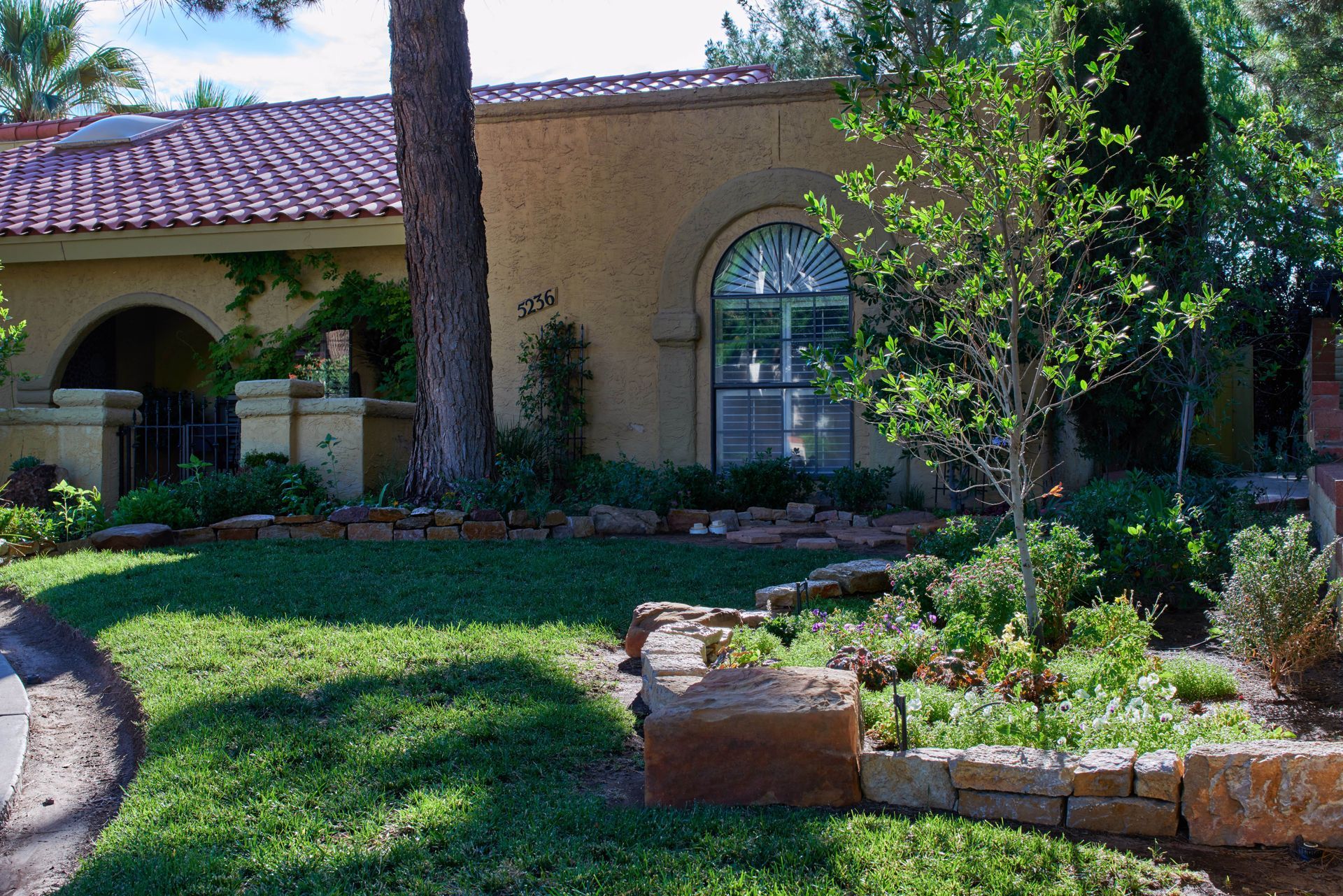 Tan stucco house with red tile roof, arched window, and landscaped yard.
