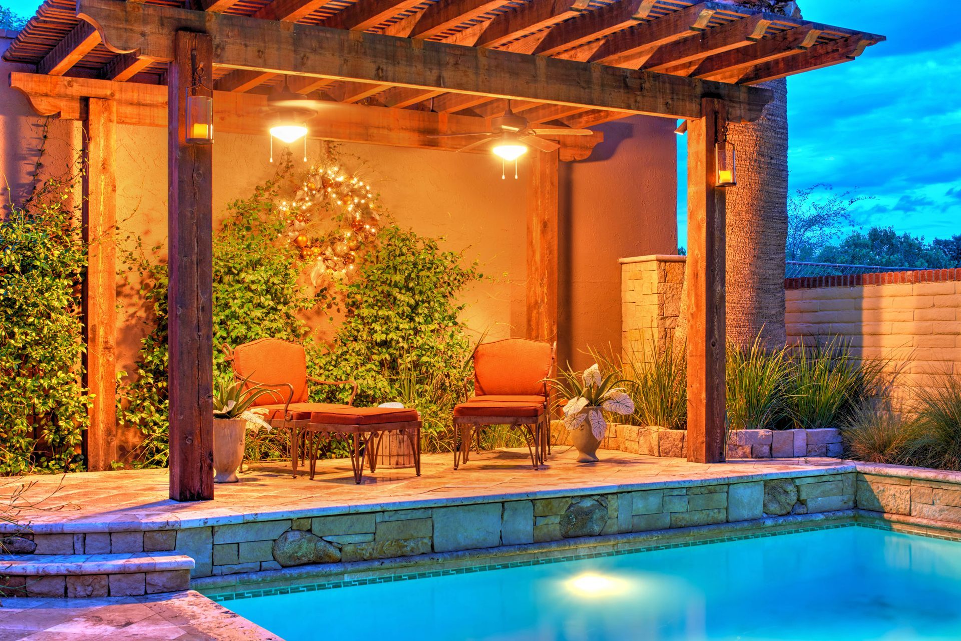 Poolside pergola with seating, overhead lighting, and lush greenery; evening ambiance.