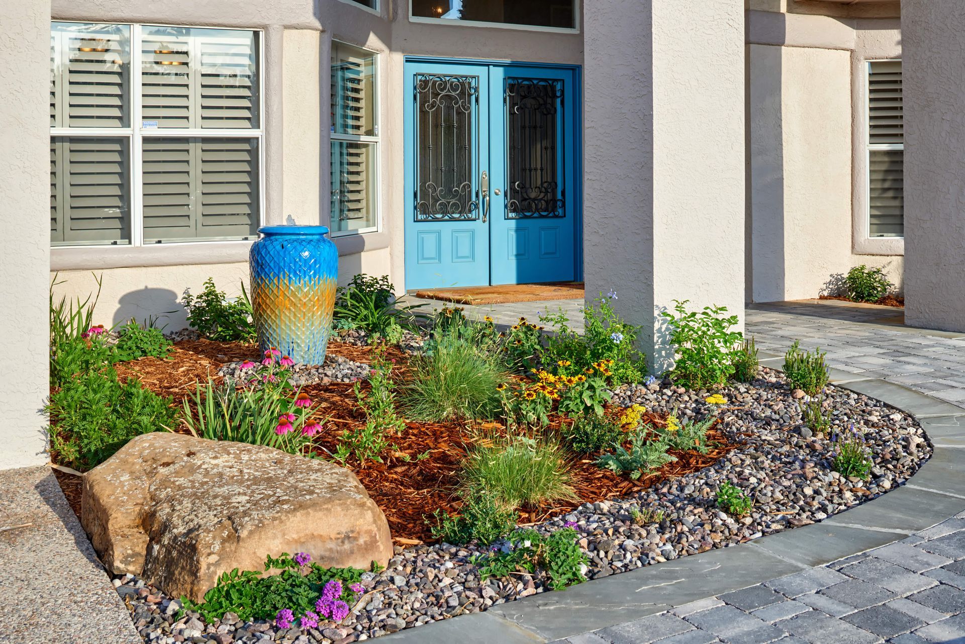 Front yard landscaping with a blue water fountain, colorful flowers, and a large rock.