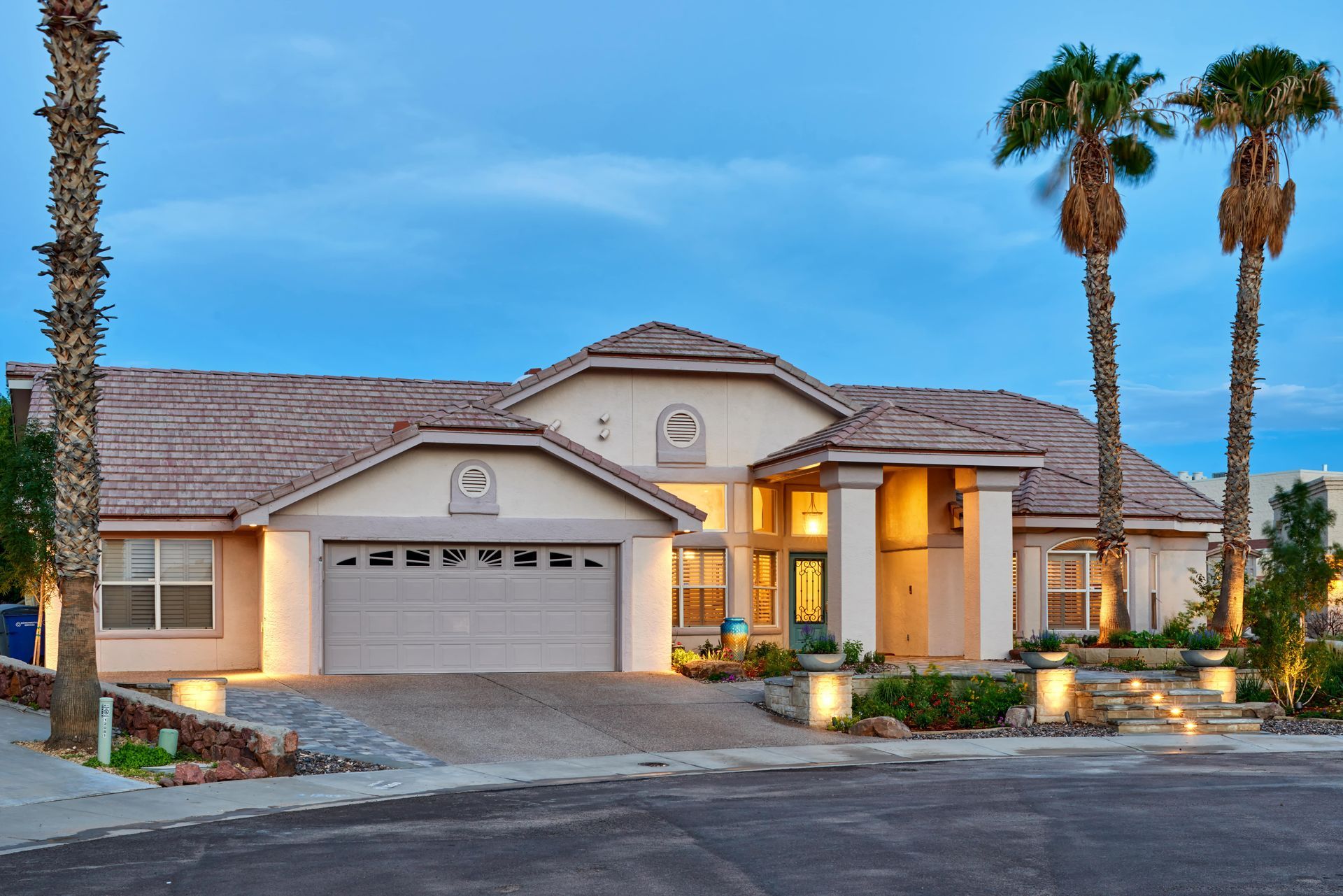 Two-story house with beige stucco exterior, palm trees, and illuminated landscaping at dusk.