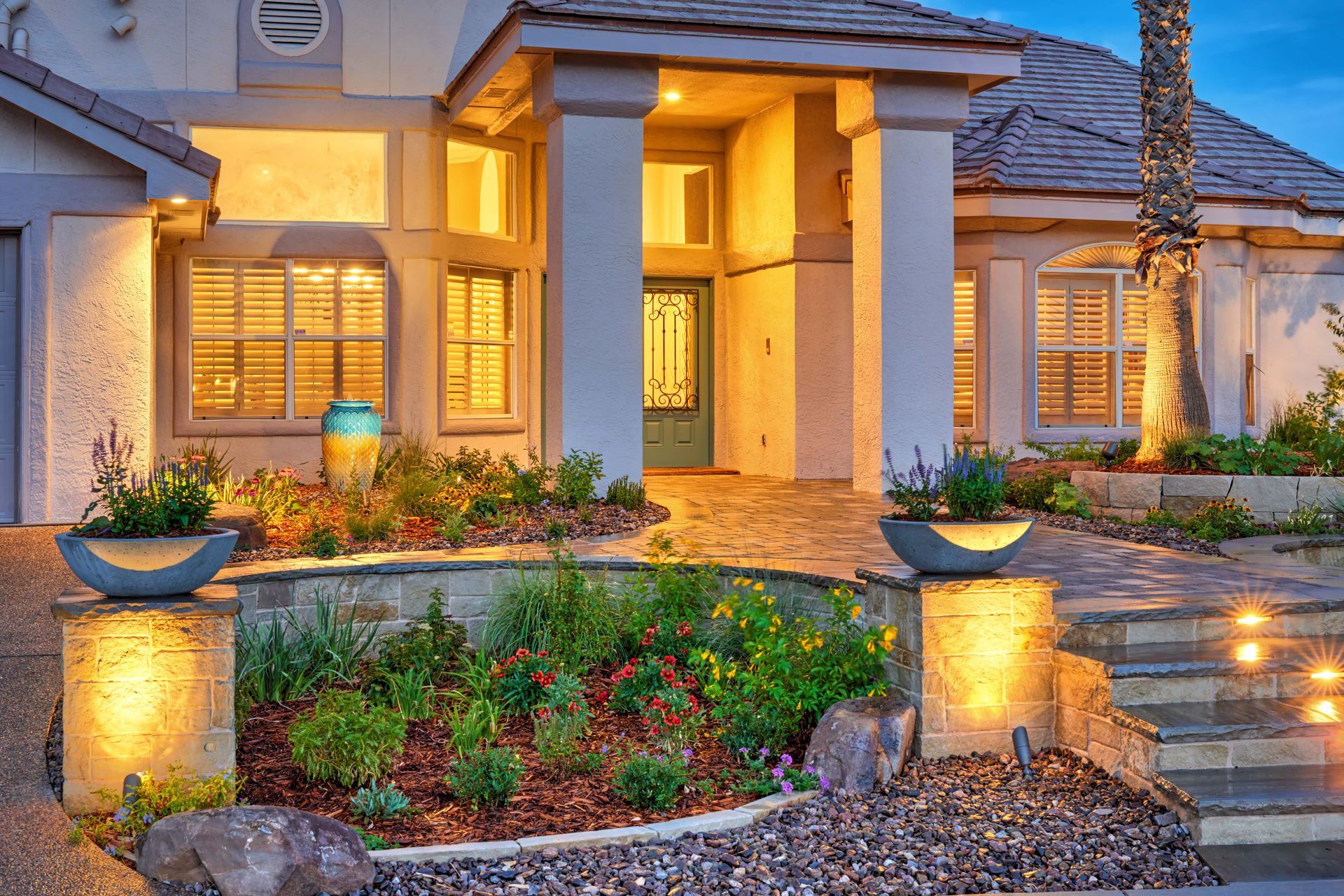 Lit-up home exterior at dusk. Features a front door, landscaping, columns, and accent lighting.