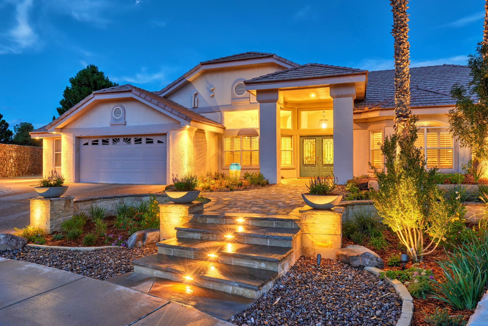 Elegant two-story house at dusk, lit entry stairs, tan stucco, arched windows, and a two-car garage.