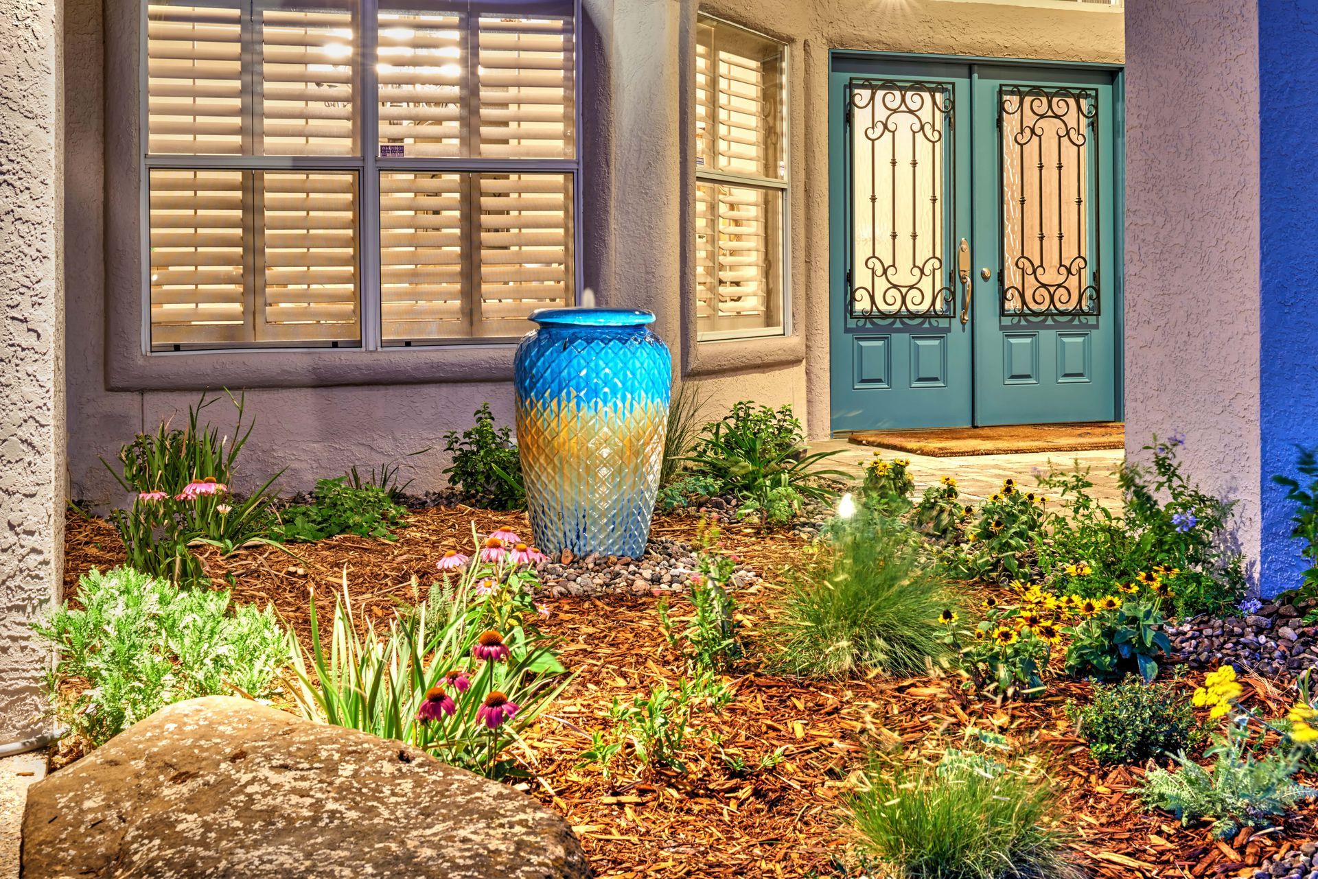 Entrance with teal doors and a blue and gold vase fountain in a flowerbed with shutters.
