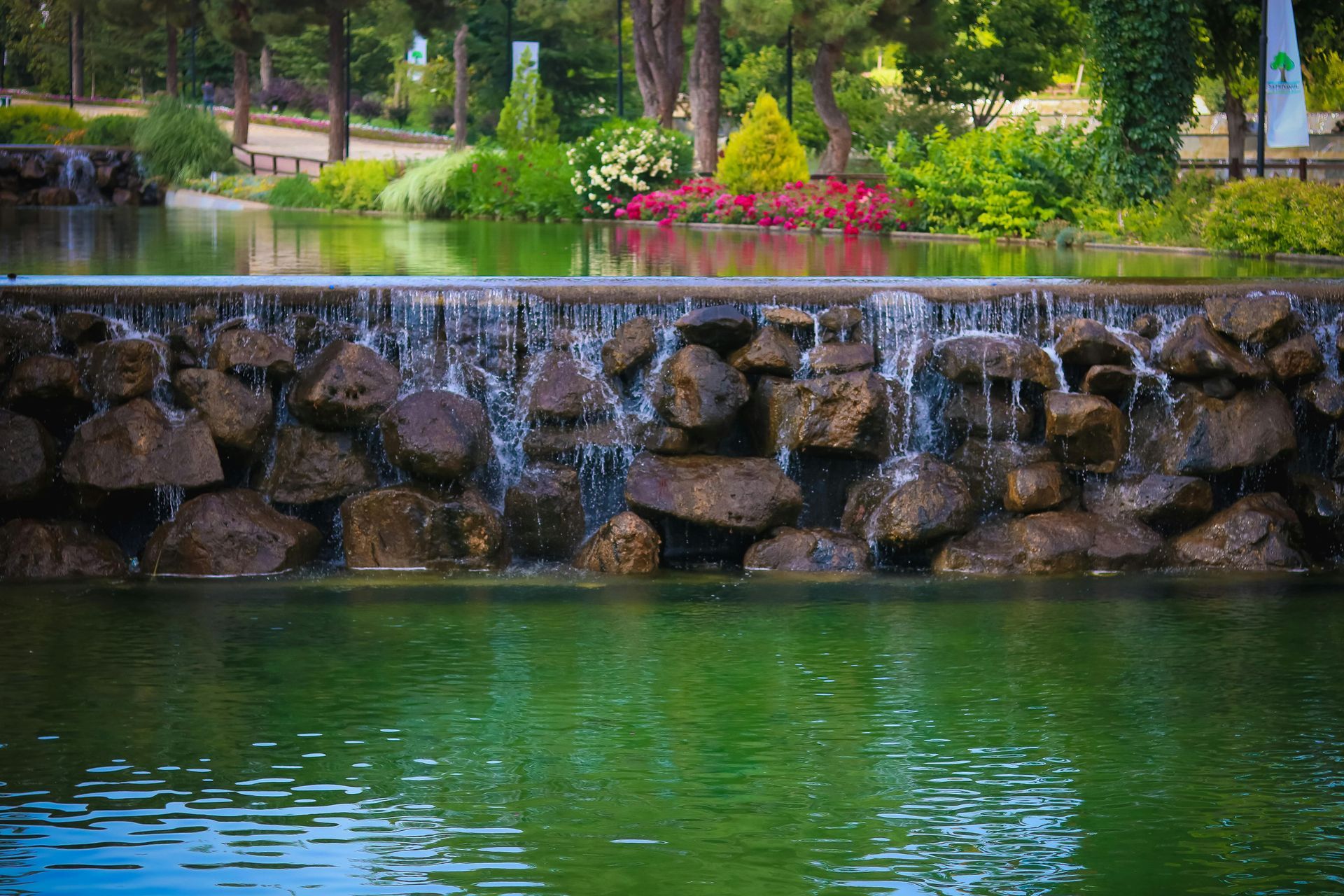 Water cascading over a rock wall into a green pond, surrounded by lush greenery and flowers.