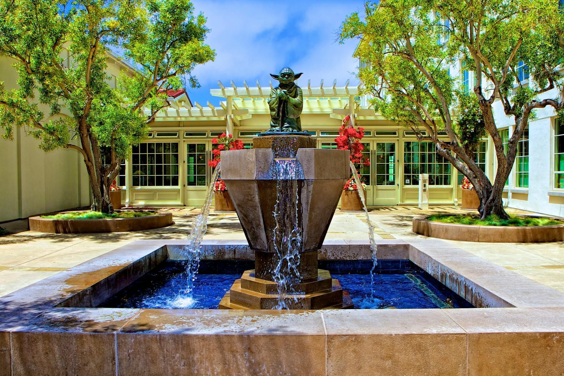Fountain with Yoda statue in courtyard, surrounded by trees and a building with windows. Water flowing.