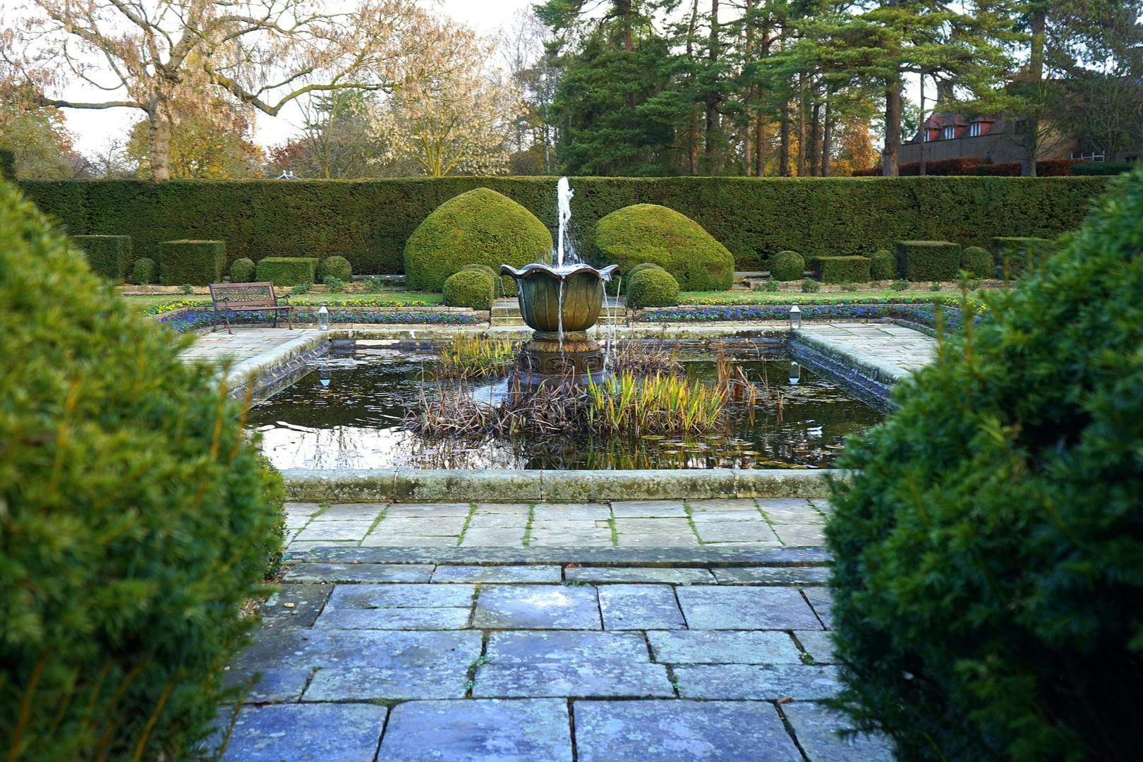 Formal garden with a fountain in a rectangular pond, bordered by manicured hedges and stone pathways.