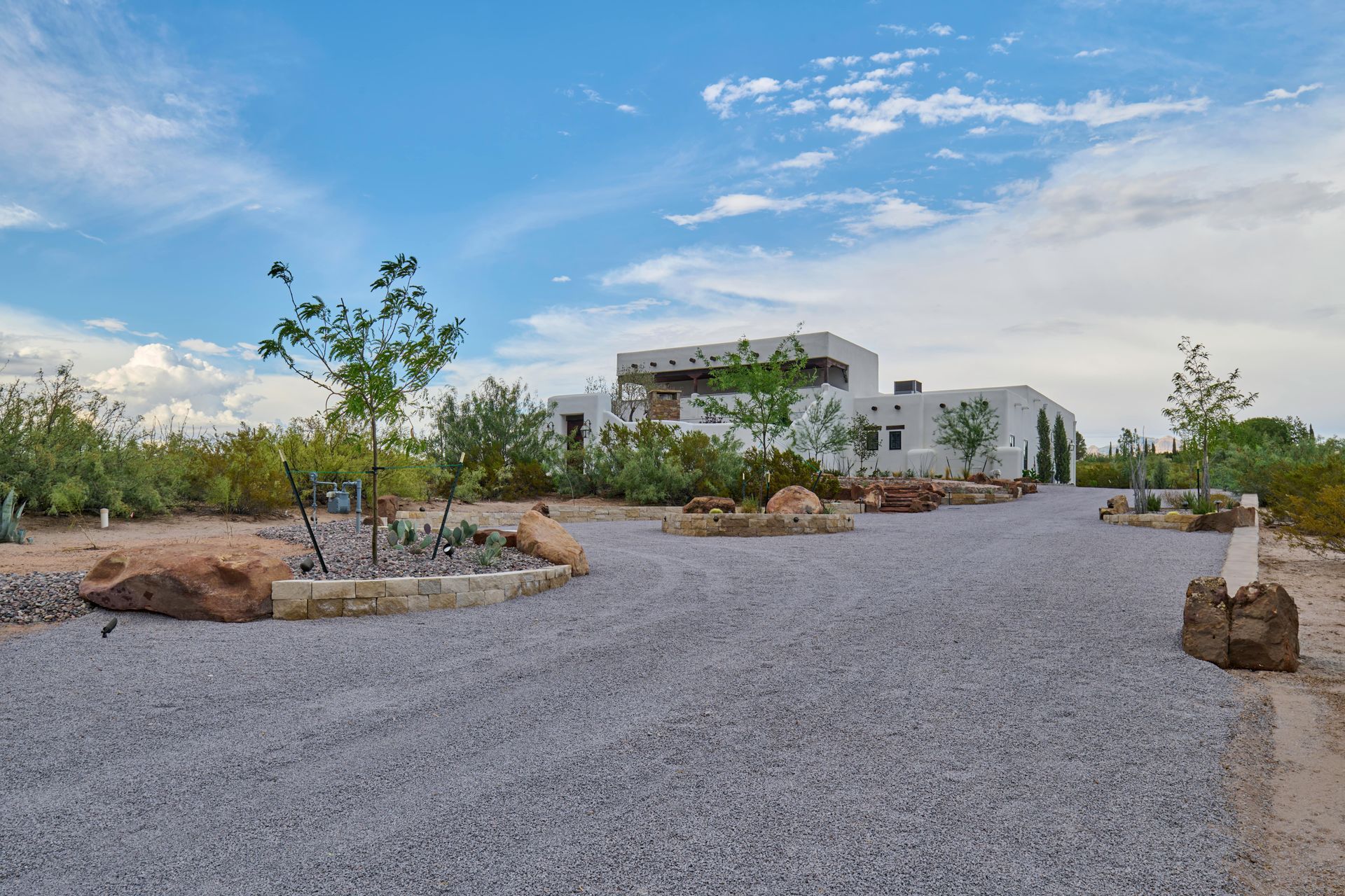 Gravel driveway leads to a white stucco house with a flat roof, surrounded by desert landscaping under a blue sky.