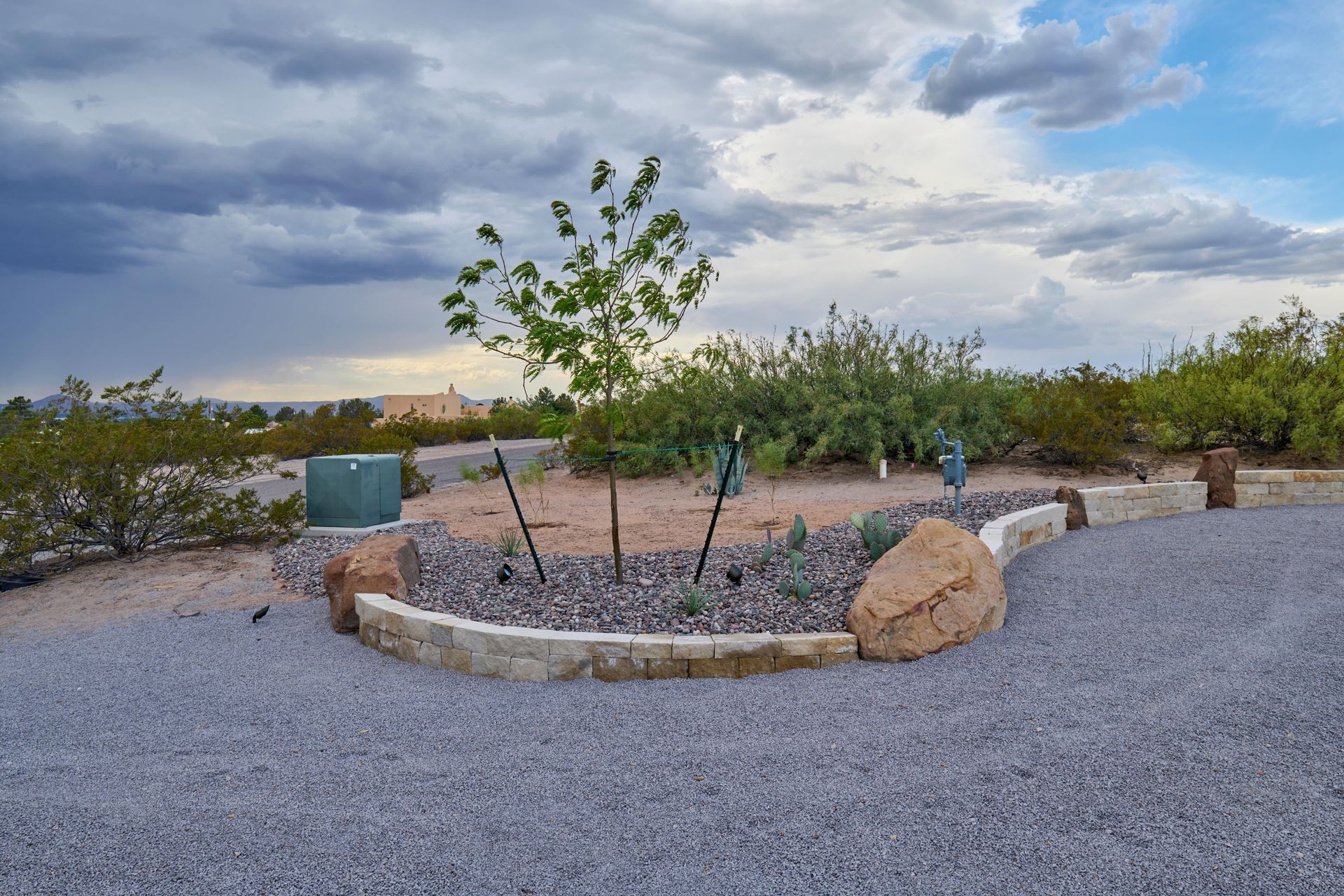 Landscaped garden bed with tree, rocks, and bushes against a cloudy sky.