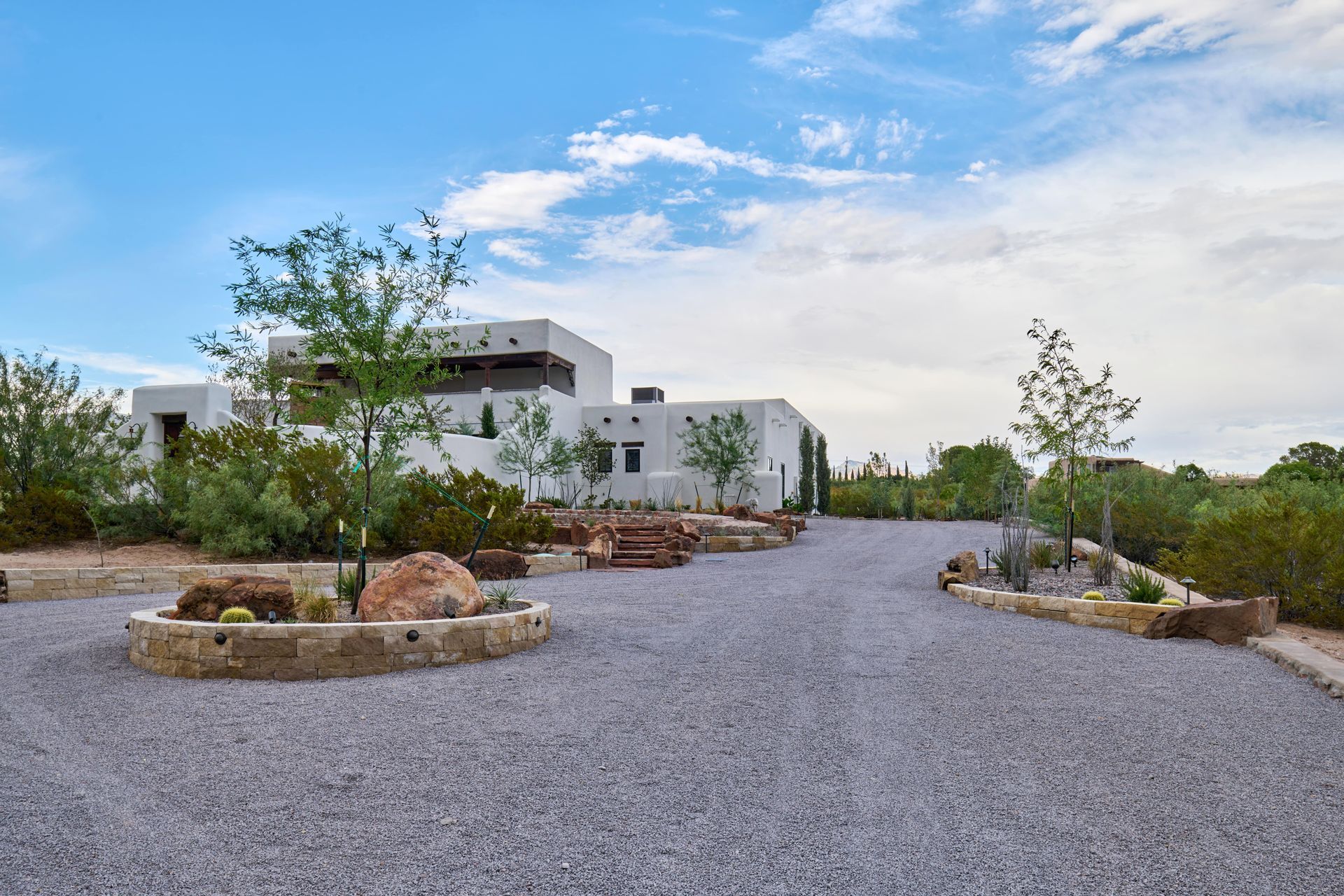 Gravel driveway leading to a white, adobe-style house with desert landscaping under a blue sky.