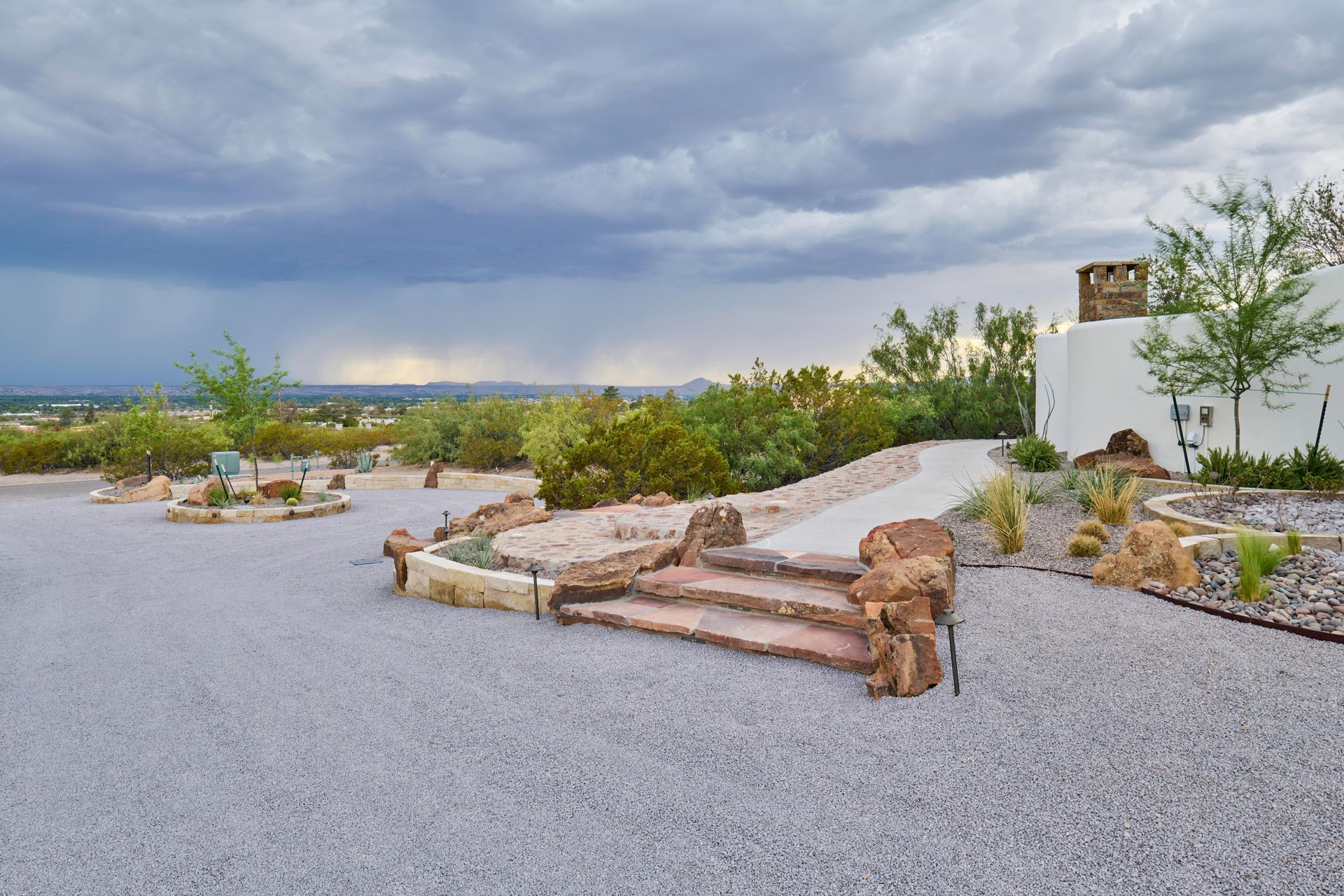 Gravel patio with stone steps and pathway, landscaping with desert plants, overlooking distant city and cloudy sky.