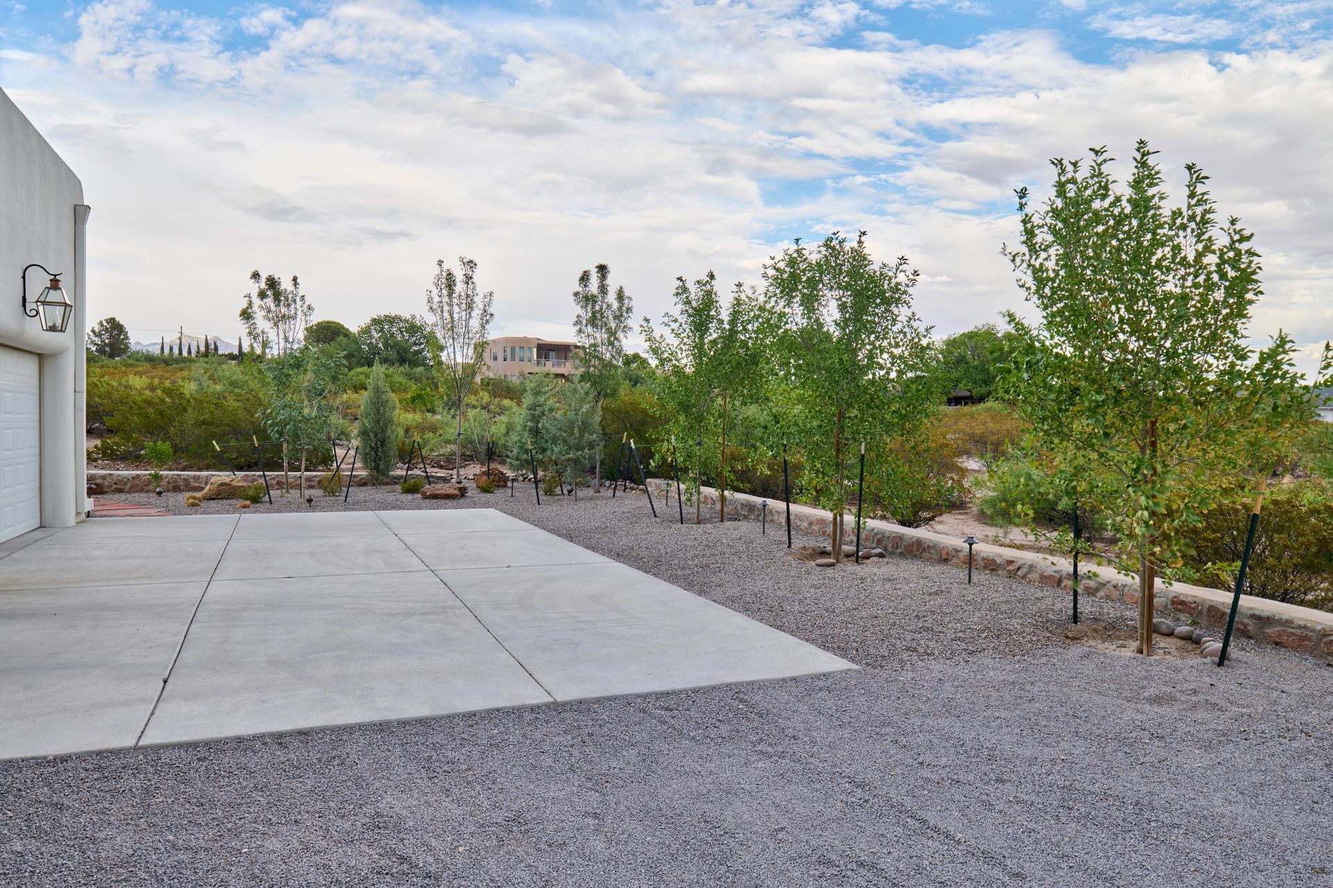 Concrete driveway next to a white building and small trees, with a gravel bed and shrubbery in the background.