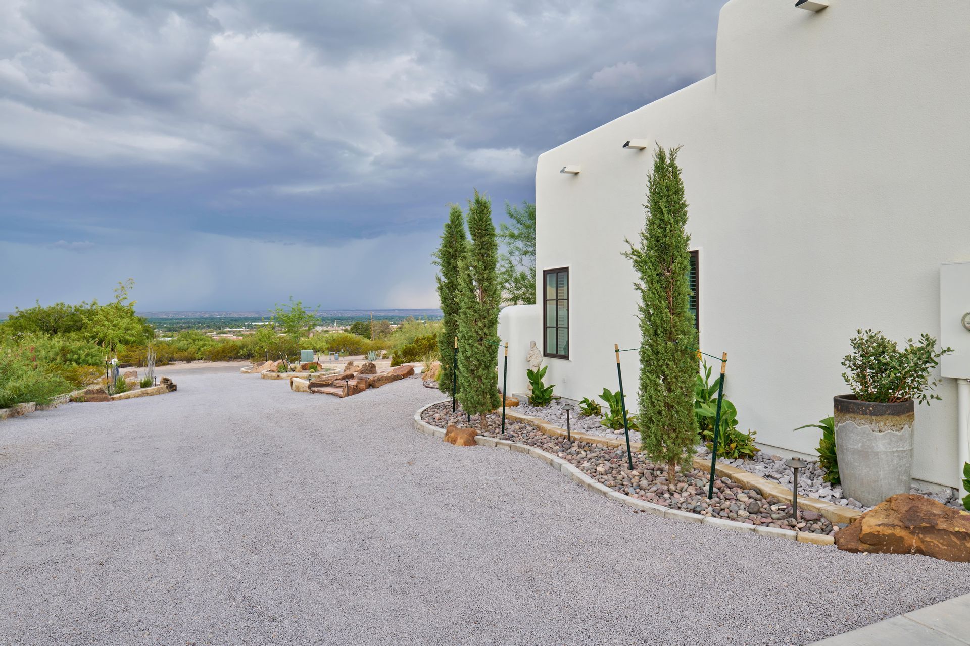 Gravel driveway leading to a white stucco building with tall thin trees and a cloudy sky.