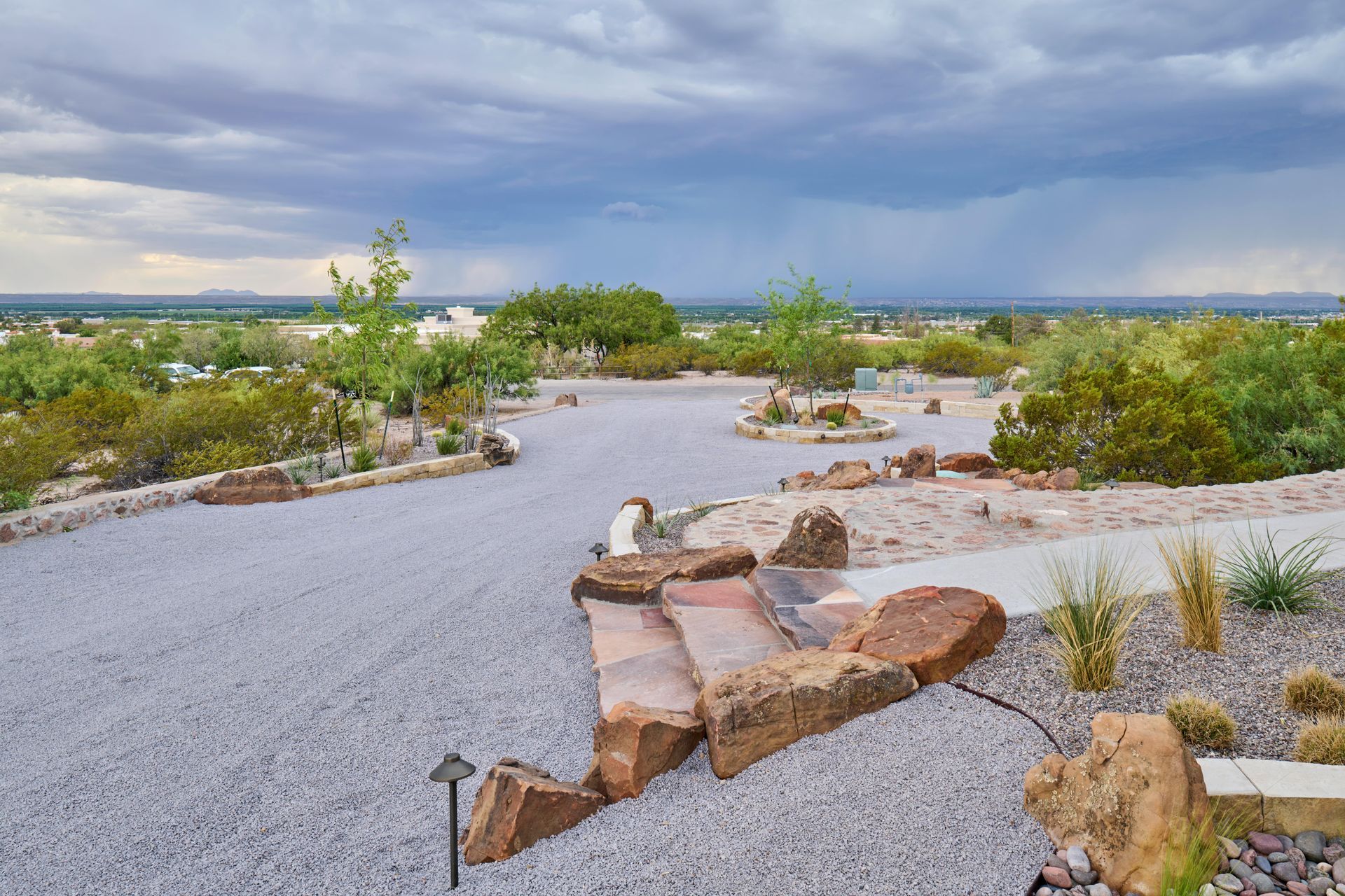 Gravel driveway and steps with large rocks, leading to a landscape with trees, under a cloudy sky.
