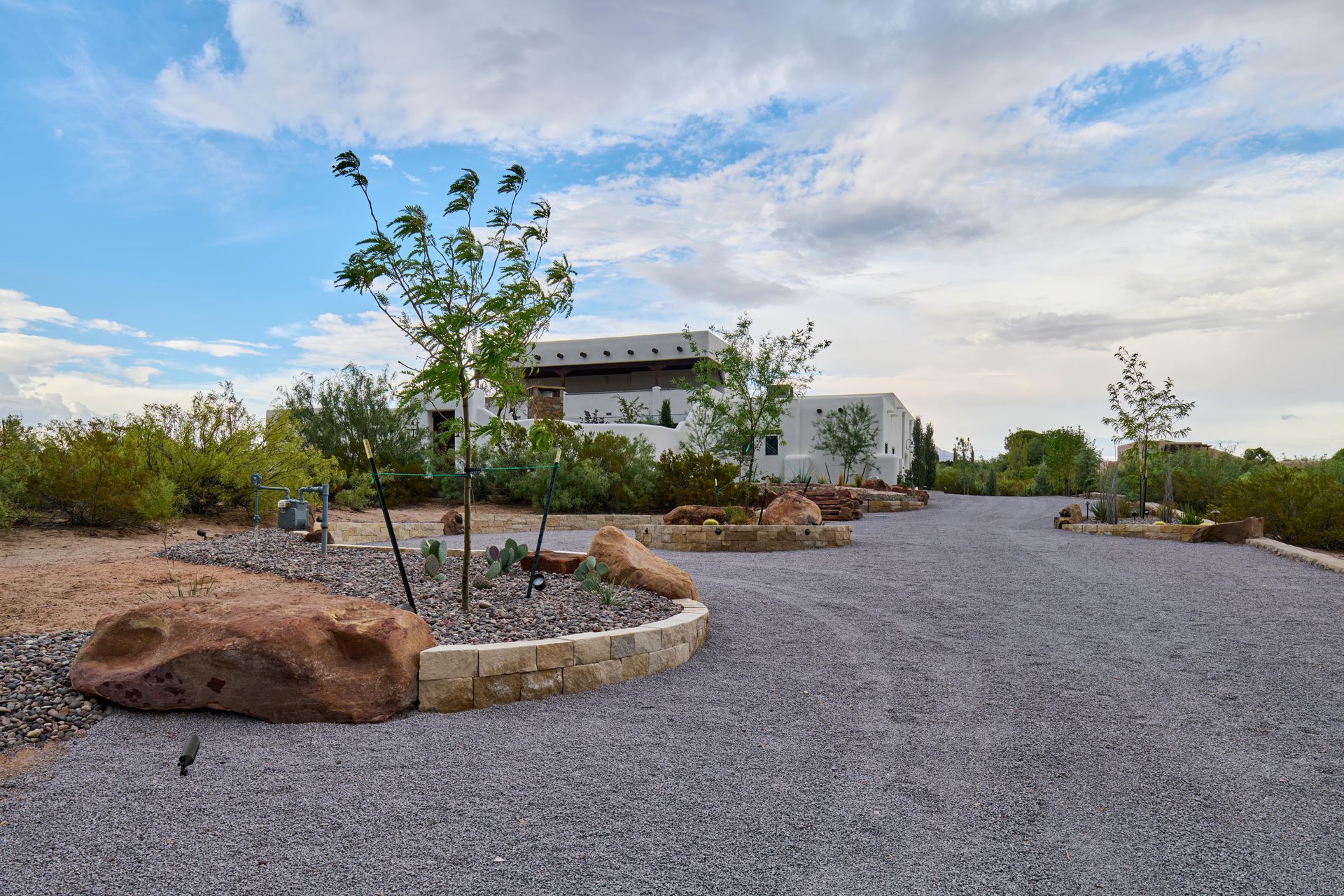 Gravel driveway leads to a white stucco building with trees and landscaping under a cloudy sky.