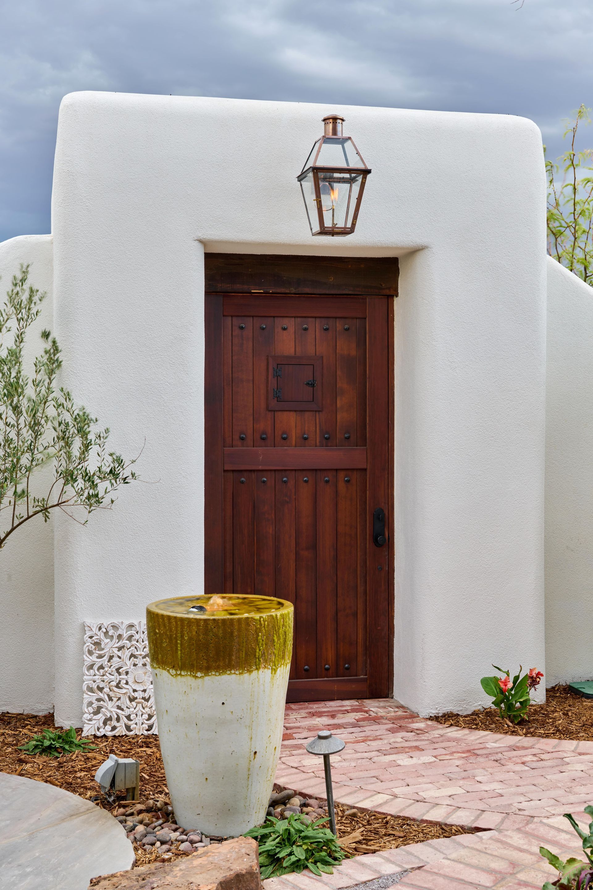 Wooden door set into a textured white wall with a bronze lantern above, and a fountain in front on a brick path.