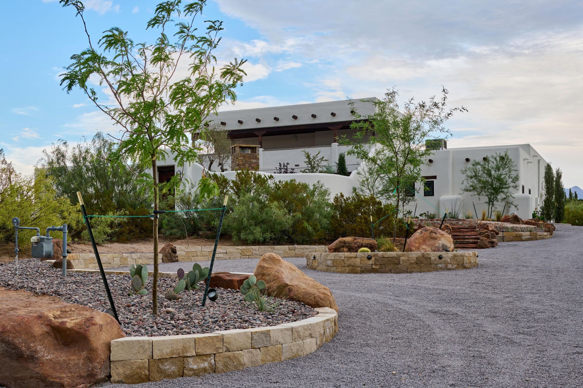 Southwestern-style building with white stucco walls, surrounded by desert landscaping and gravel driveway. Blue sky.