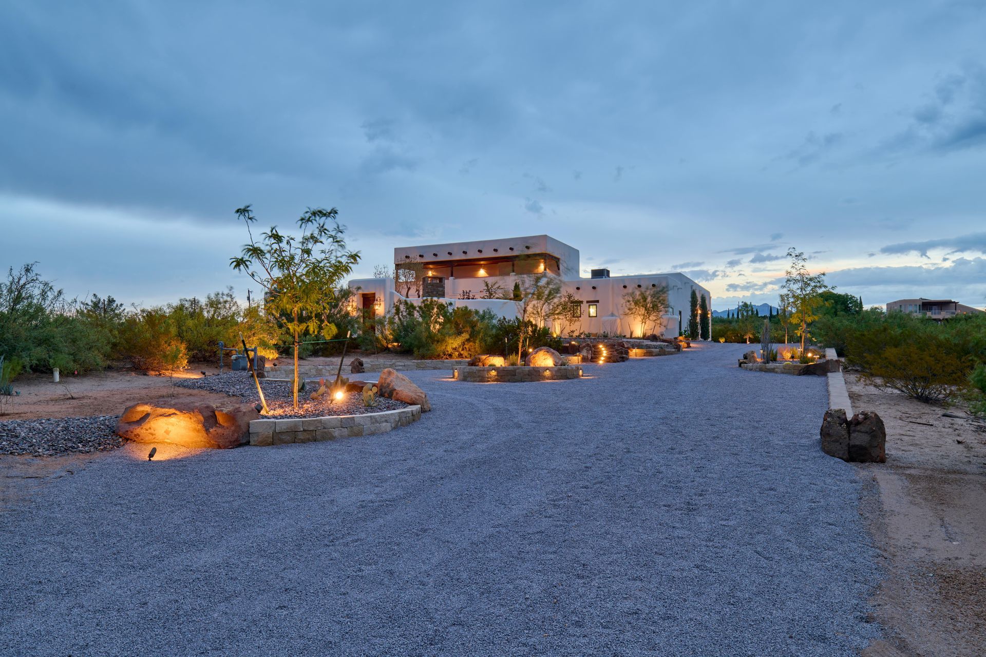 Gravel driveway leads to a Southwestern-style home at dusk, lit by exterior lights, against a cloudy sky.