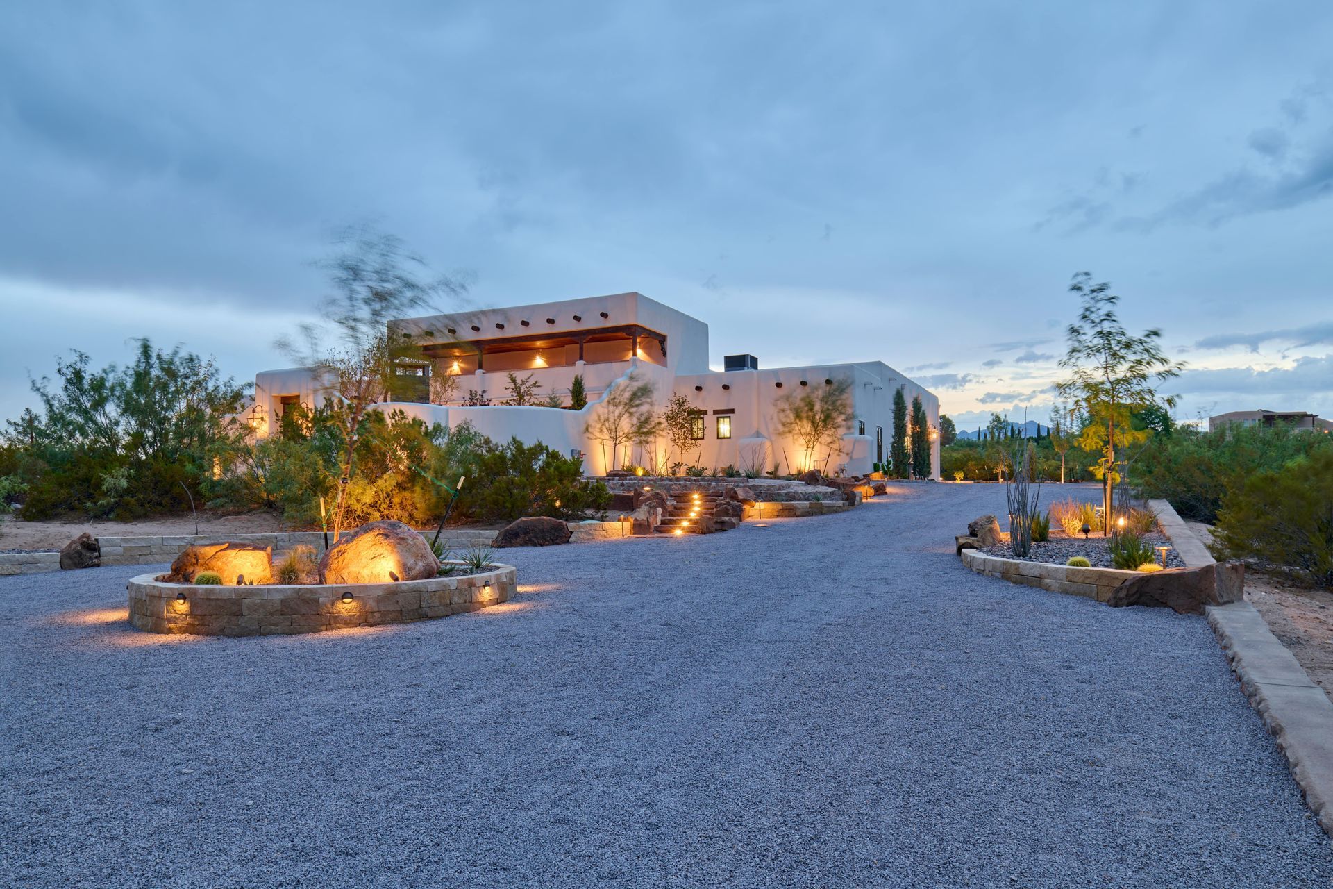 Gravel driveway leads to a Southwestern-style house at dusk. Soft lights illuminate landscaping.