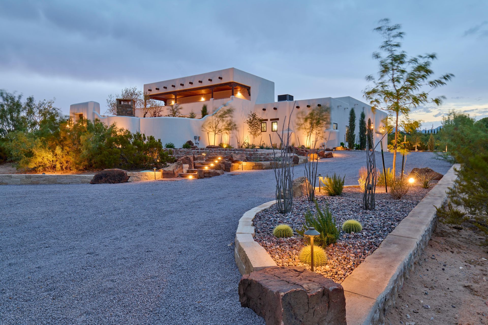 White stucco home with illuminated landscaping and gravel driveway at dusk.