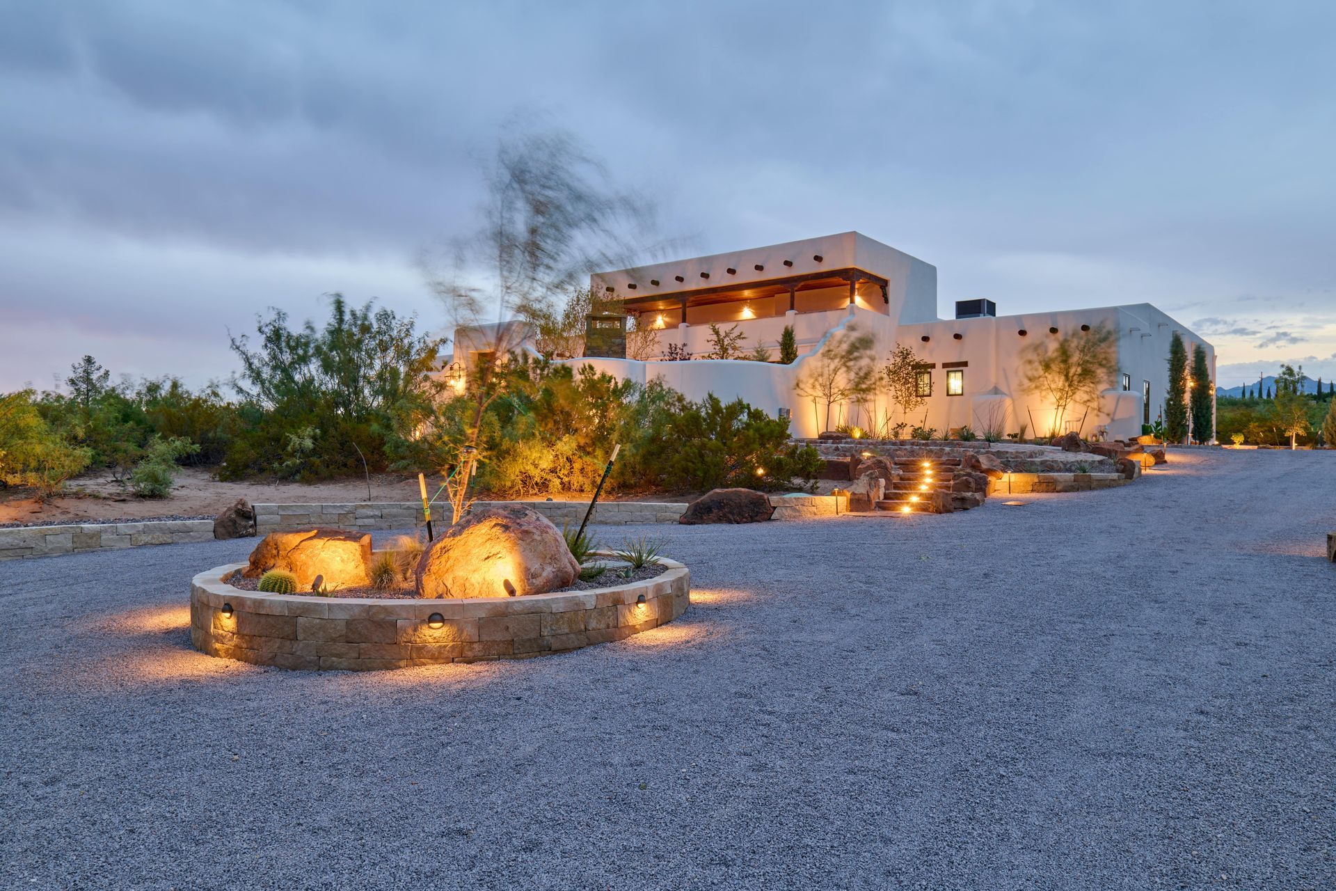 Desert home at dusk, exterior illuminated with warm lights, gravel driveway, and surrounding desert vegetation.