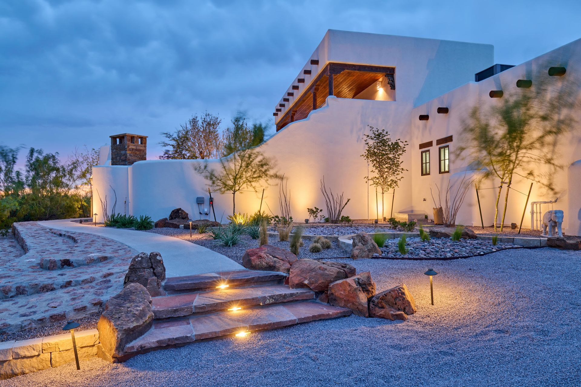 A southwestern-style building with white stucco walls, lit pathways, and a gravel yard at dusk.