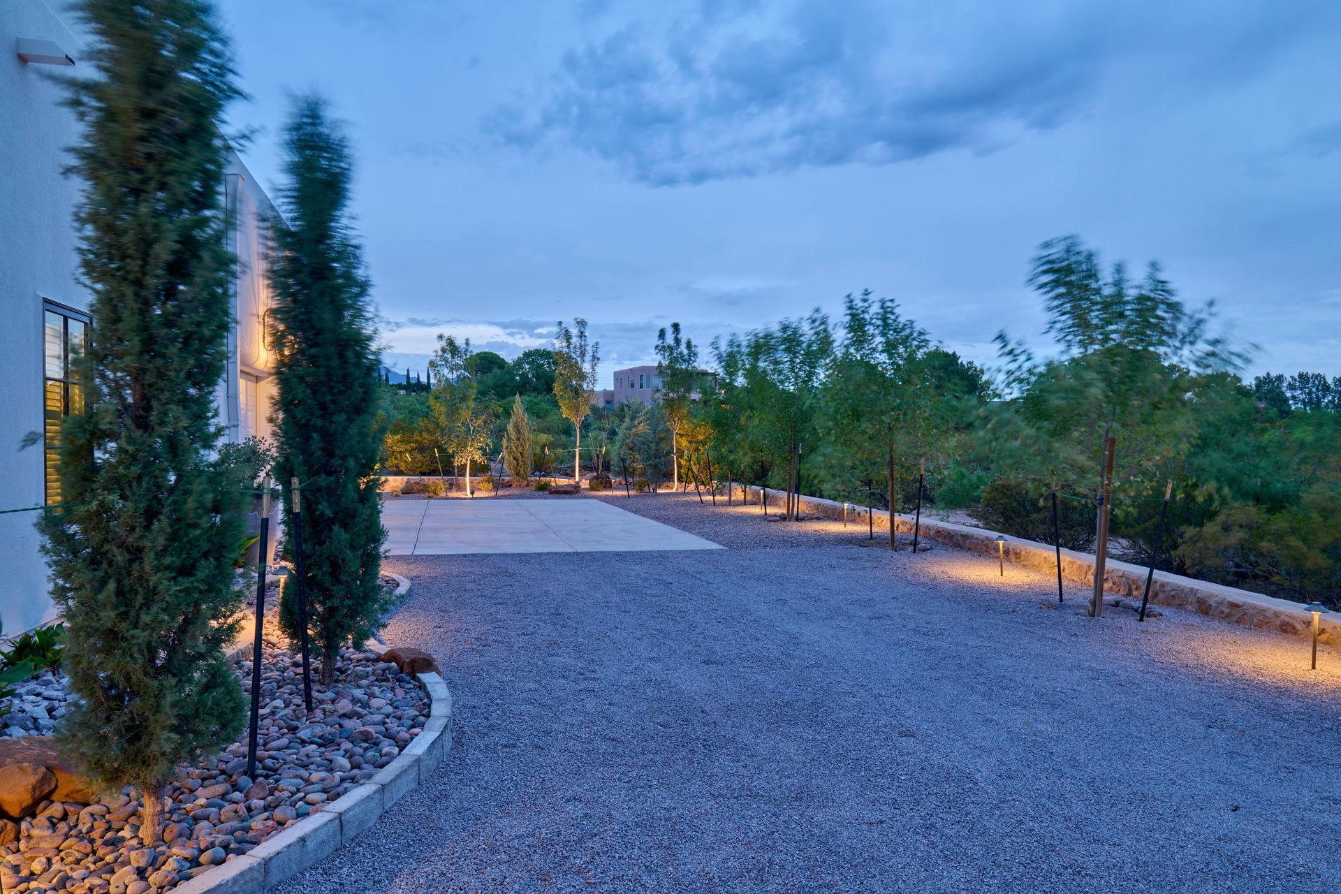 Gravel driveway lit by outdoor lights, tall trees on left and lining the perimeter at dusk.
