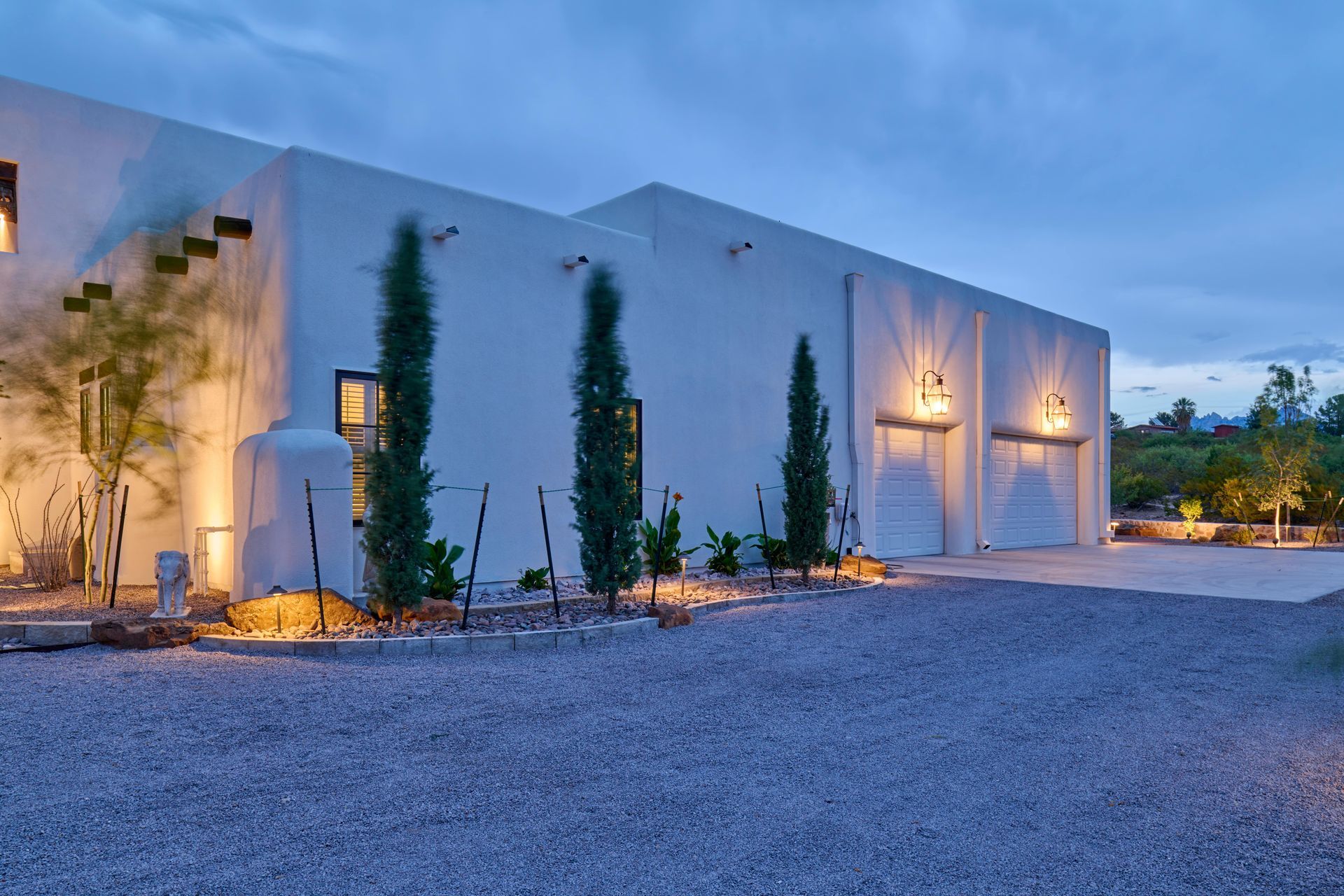 White stucco building with a gravel driveway, illuminated by outdoor lights at dusk, with cypress trees.