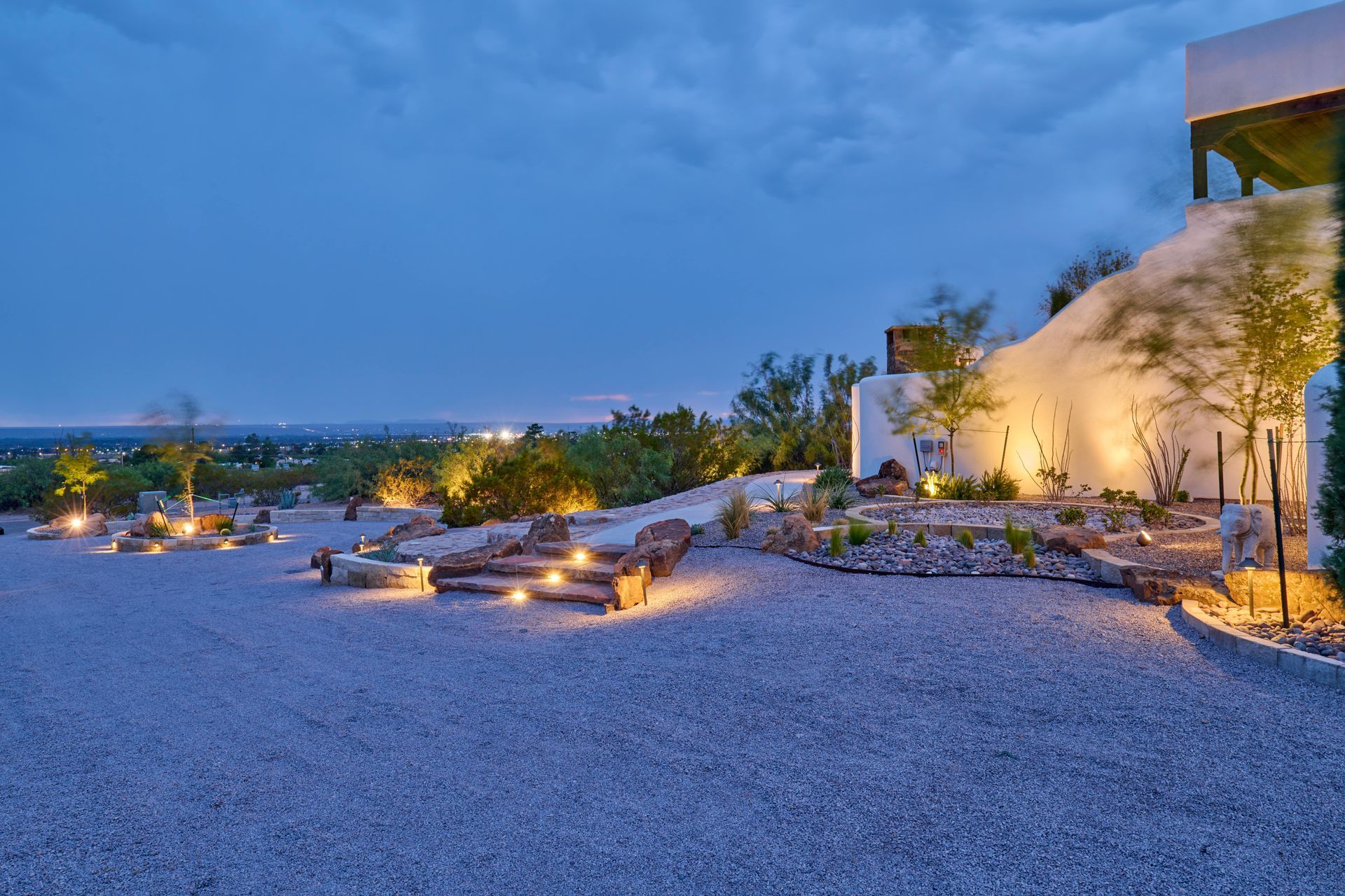 Gravel driveway at dusk, illuminated landscaping with lights, white stucco wall, view of distant city lights.