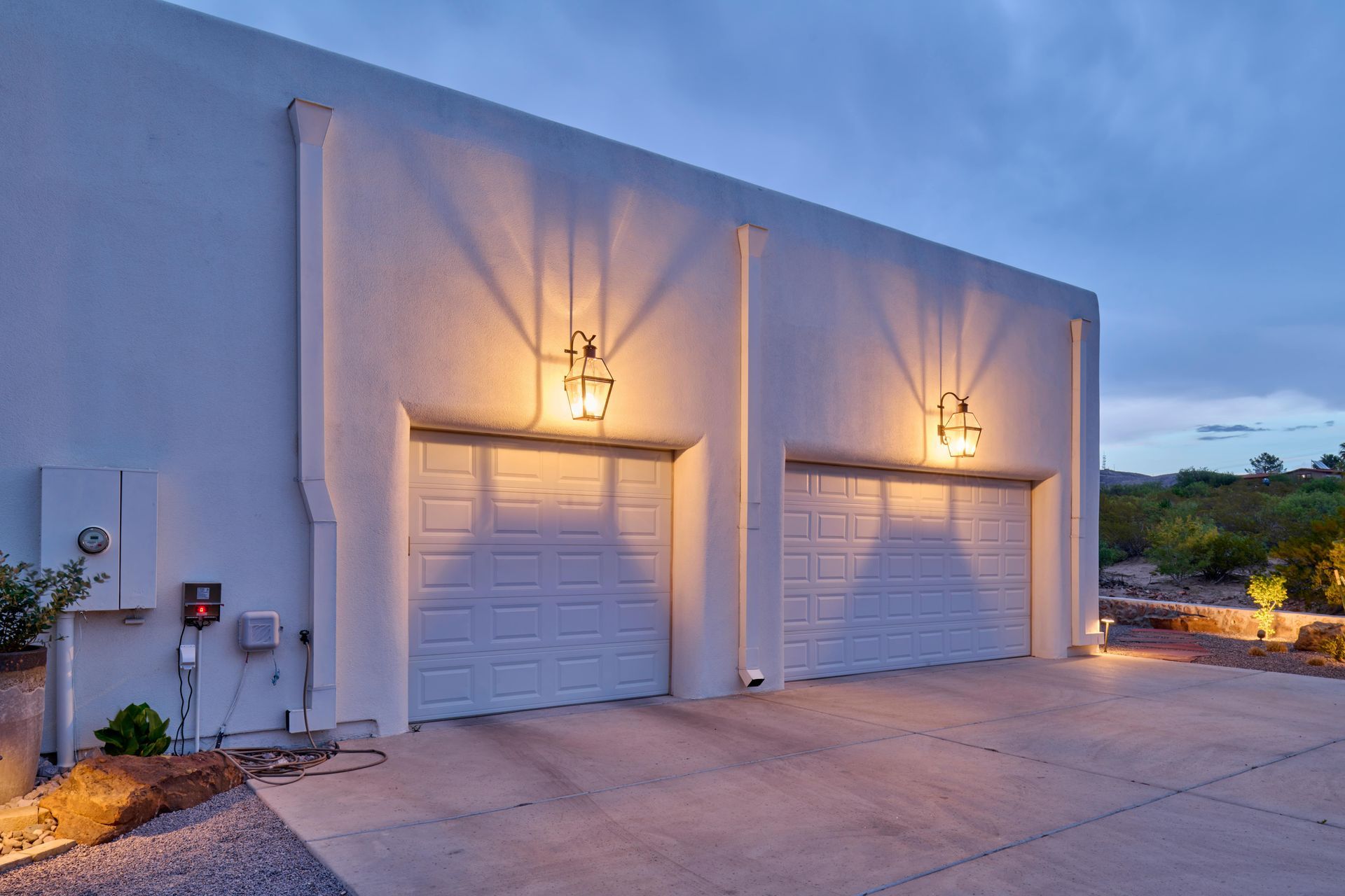 Two white garage doors on a stucco building illuminated by wall sconces at dusk.