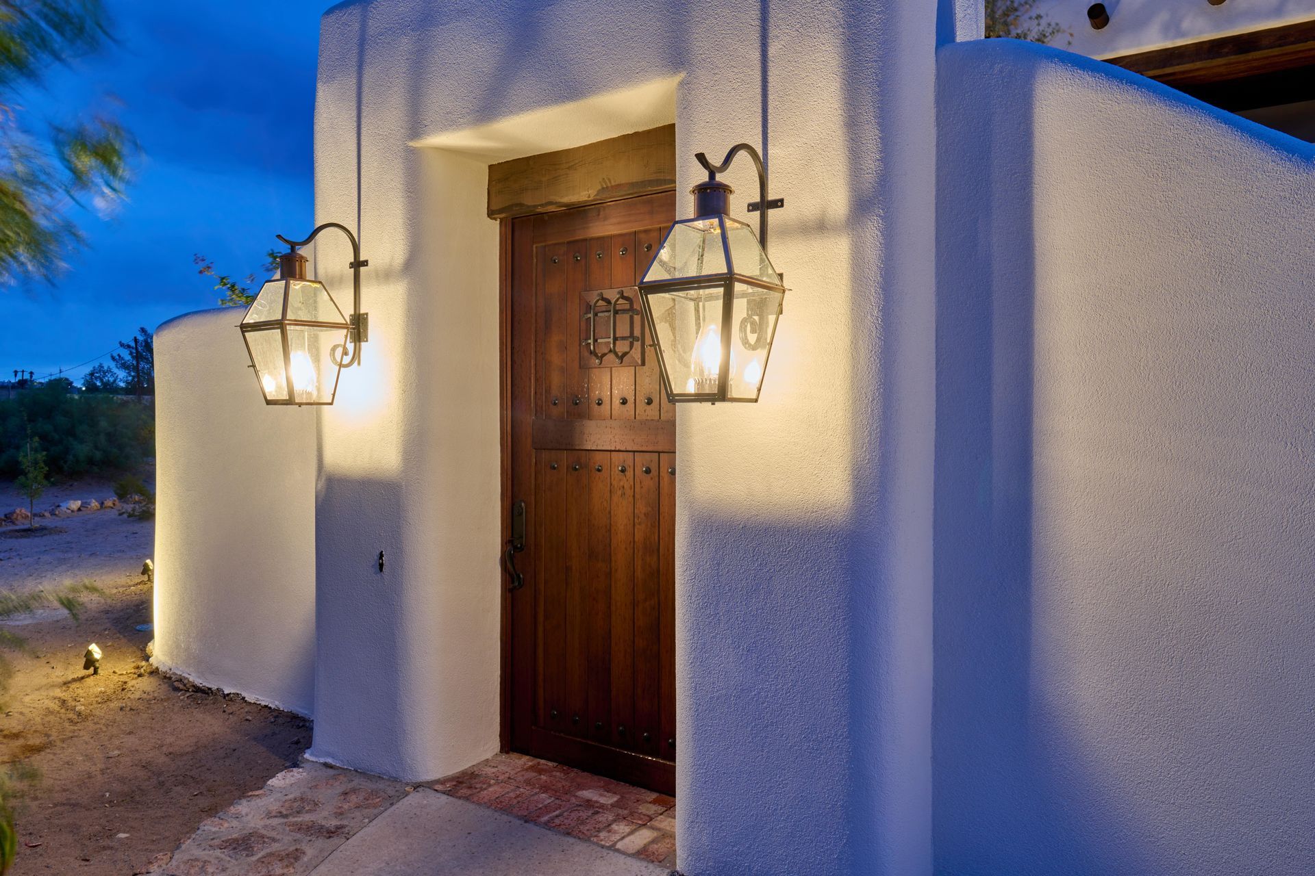 Exterior of a house at dusk, with wooden door and lantern style wall lights. White stucco walls.