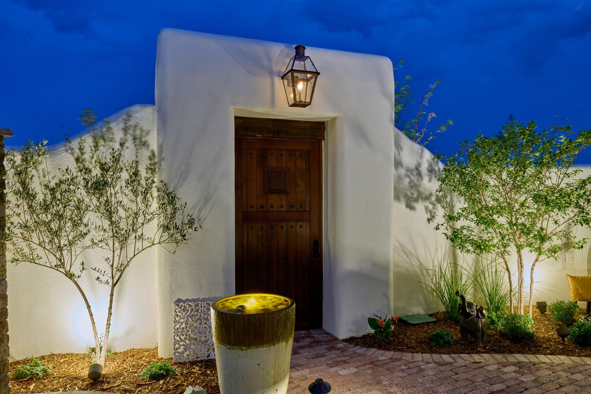 Adobe entry with a wooden door, surrounded by white stucco walls and illuminated by a lantern, at dusk.