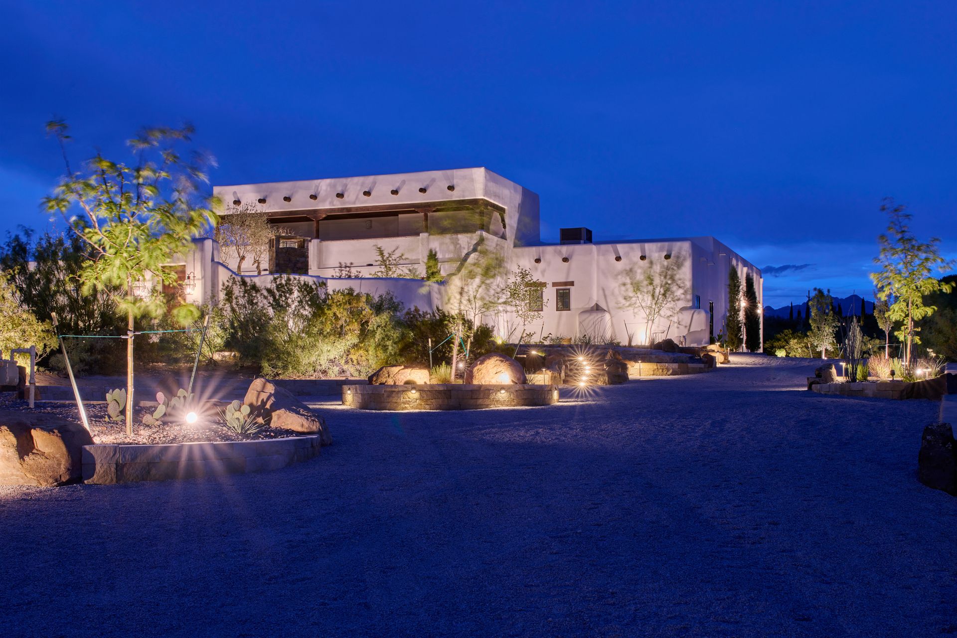 A white adobe-style house at dusk with outdoor lighting and a gravel driveway.