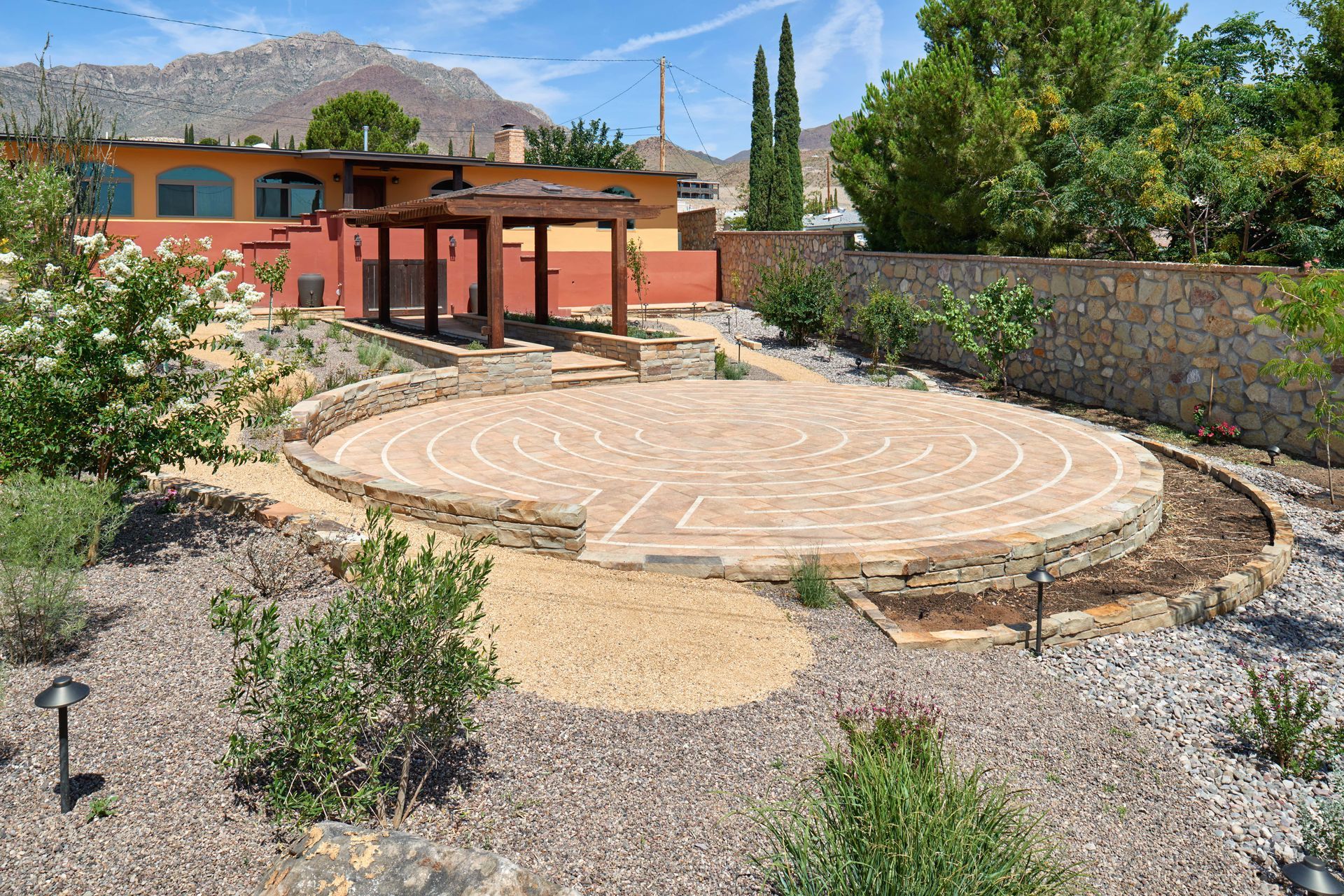 Circular labyrinth in a garden with stone pathways and a wooden pergola, under a blue sky.