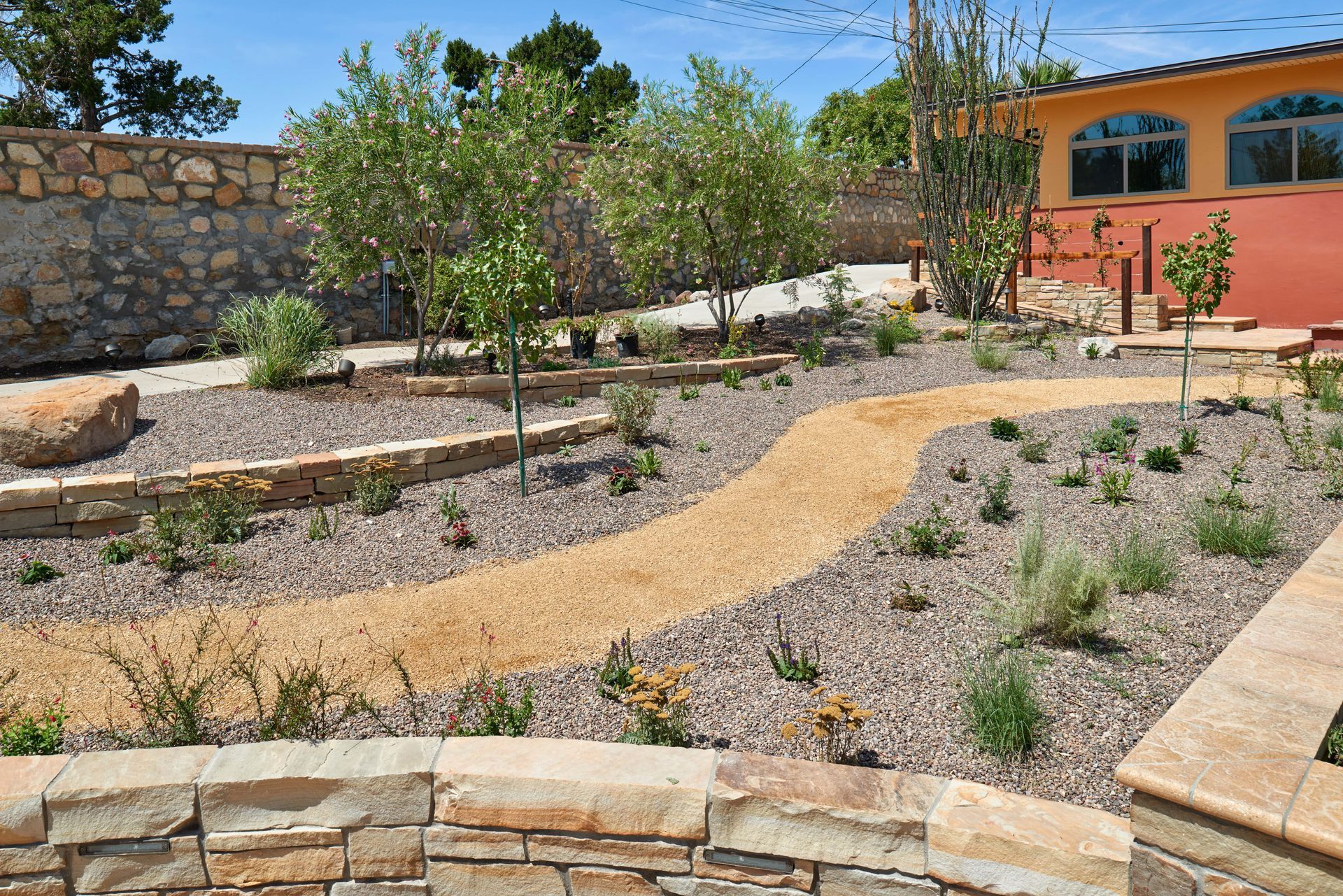 Gravel pathways wind through a landscaped garden with trees, plants, and a stone wall.