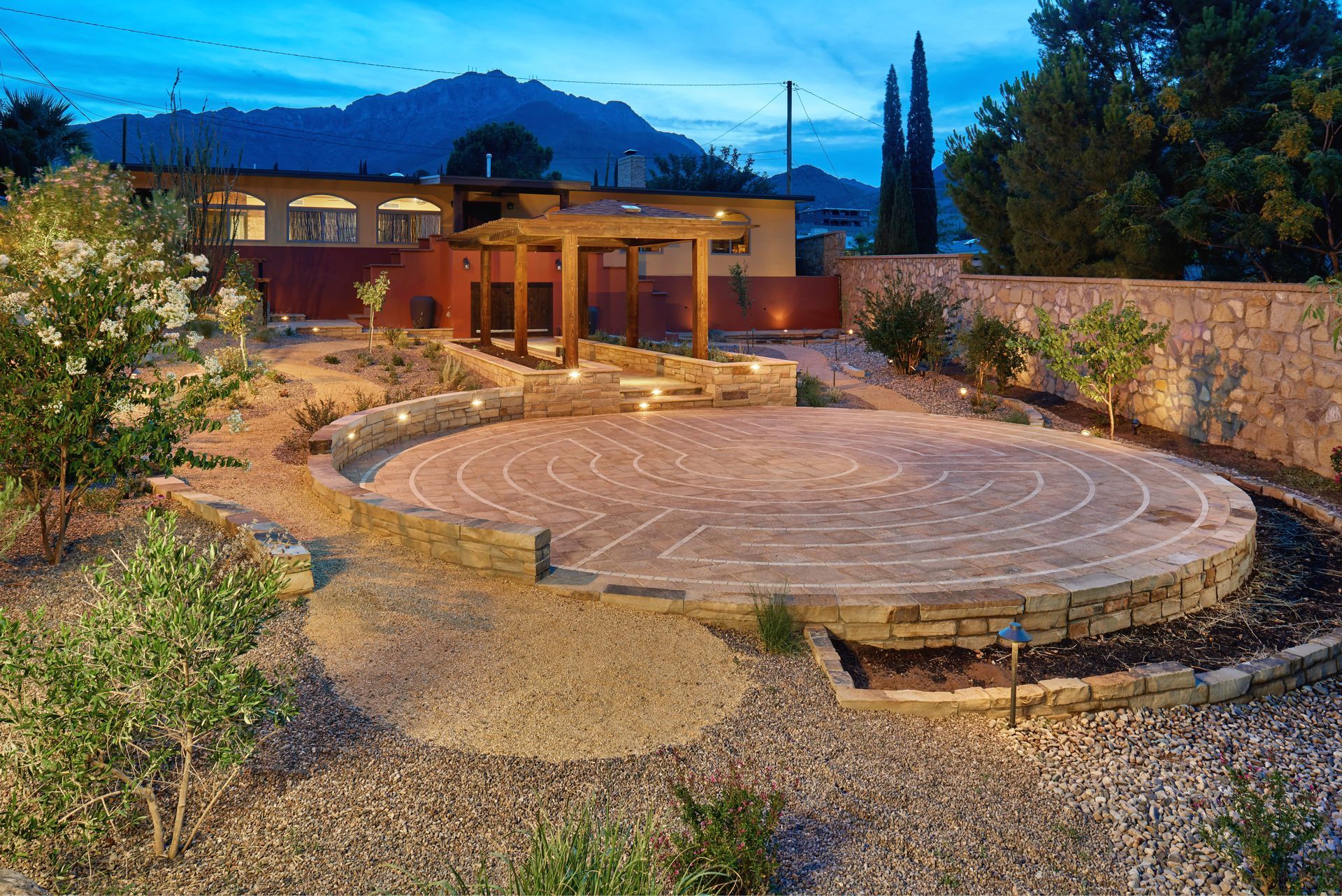 Stone labyrinth in a desert landscape, lit at dusk. Building and mountain visible in the background.