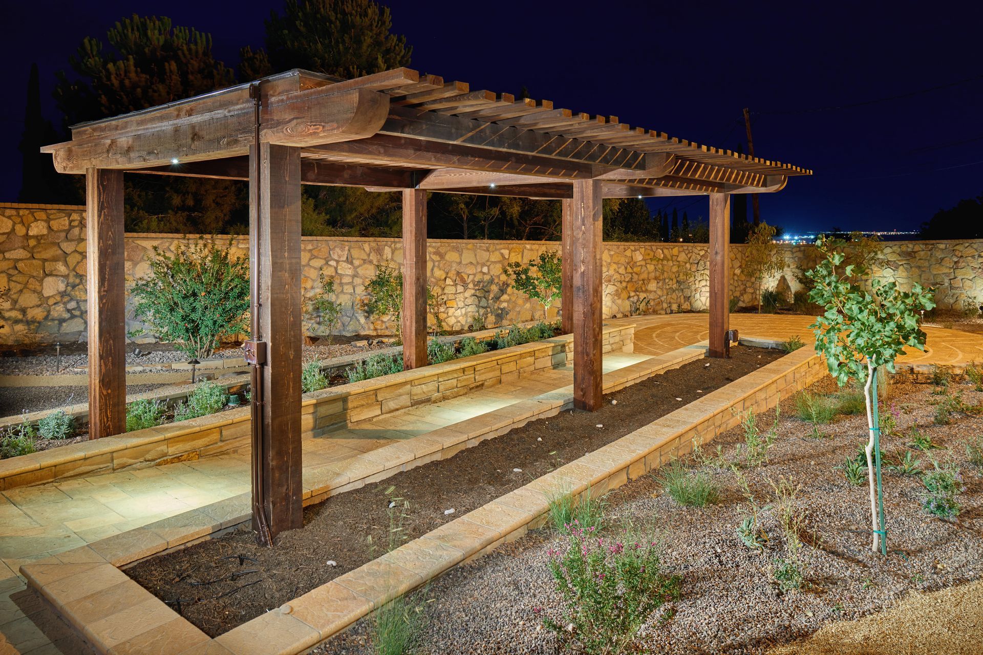 Wooden pergola in a garden at night, lit by spotlights, with a stone wall backdrop and small trees.