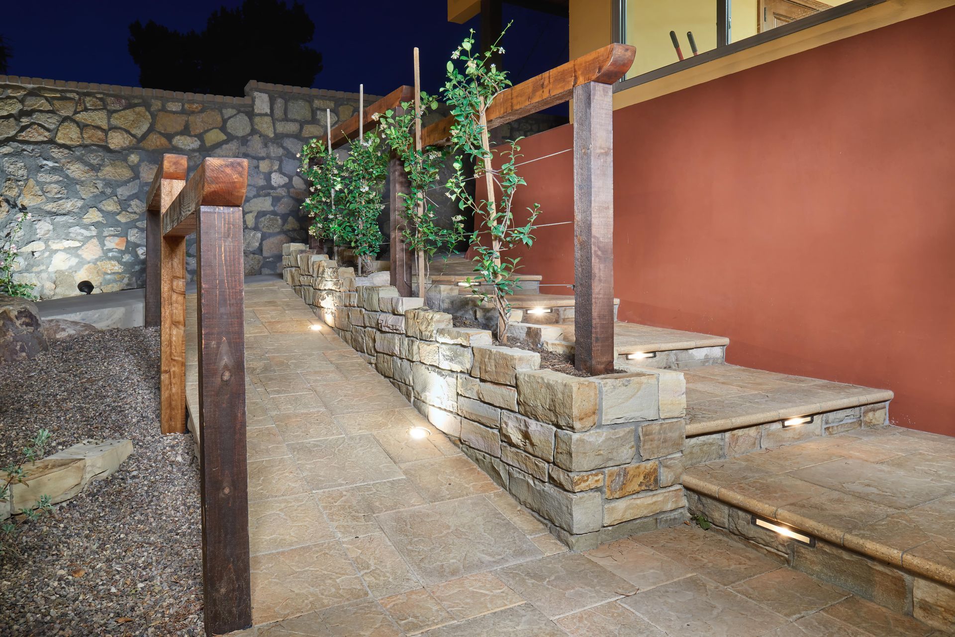 Stone walkway and steps with wooden trellis supporting climbing plants, lit by spotlights.
