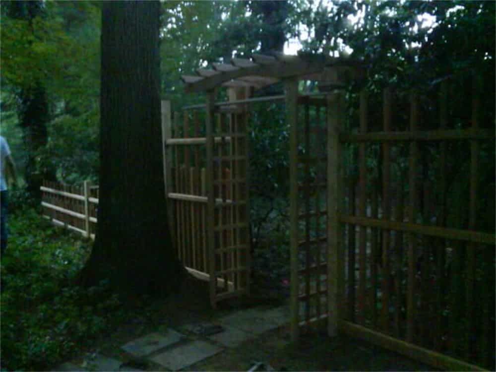 Wooden garden gate with a trellis and surrounding fence in a wooded area.
