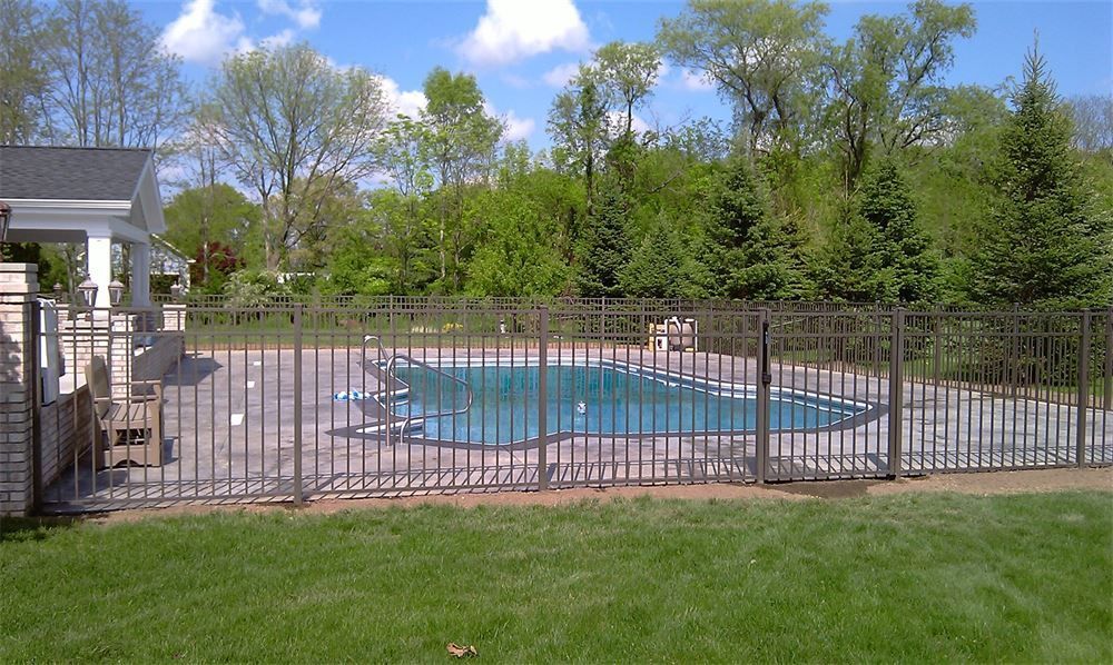 Fenced-in pool area with a house on the left and trees in the background. Green grass in the foreground.